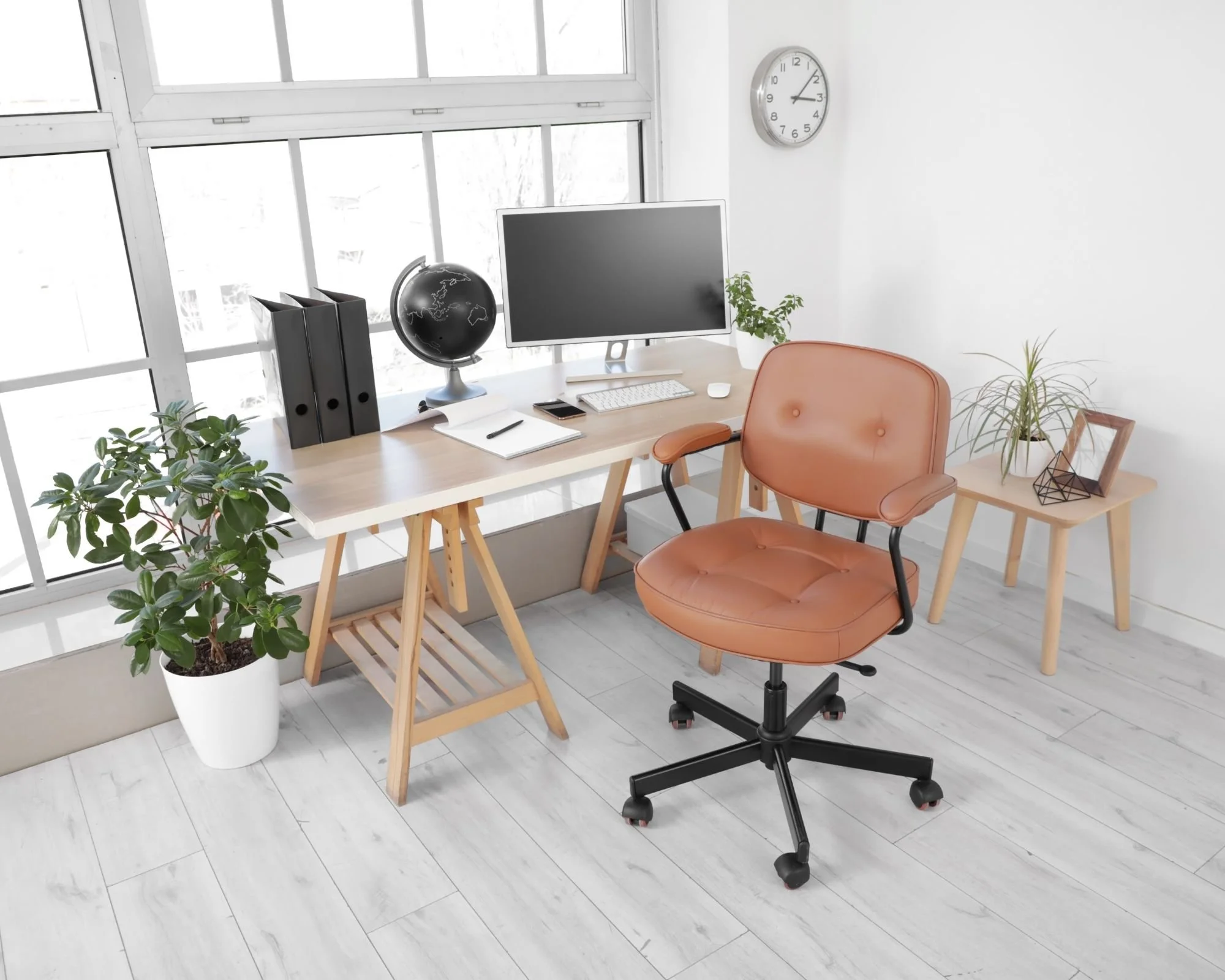 Modern office space with a wooden desk, a computer monitor, keyboard, mouse, black globe, and three black binders. There is a tan office chair, a side table with potted plants, a wall clock, and large windows letting in natural light.