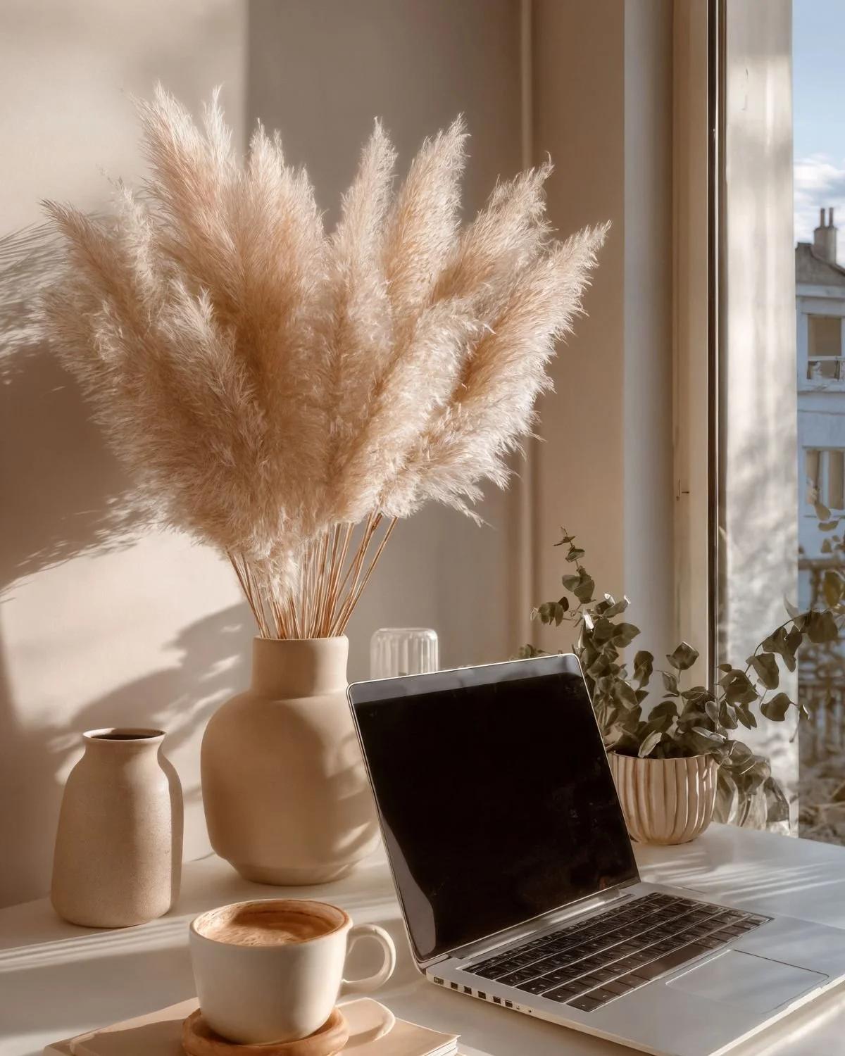 A cozy desk scene near a window with a large vase of pampas grass, a smaller vase, a cup of coffee on a saucer, a laptop, and a potted plant, all bathed in warm sunlight.