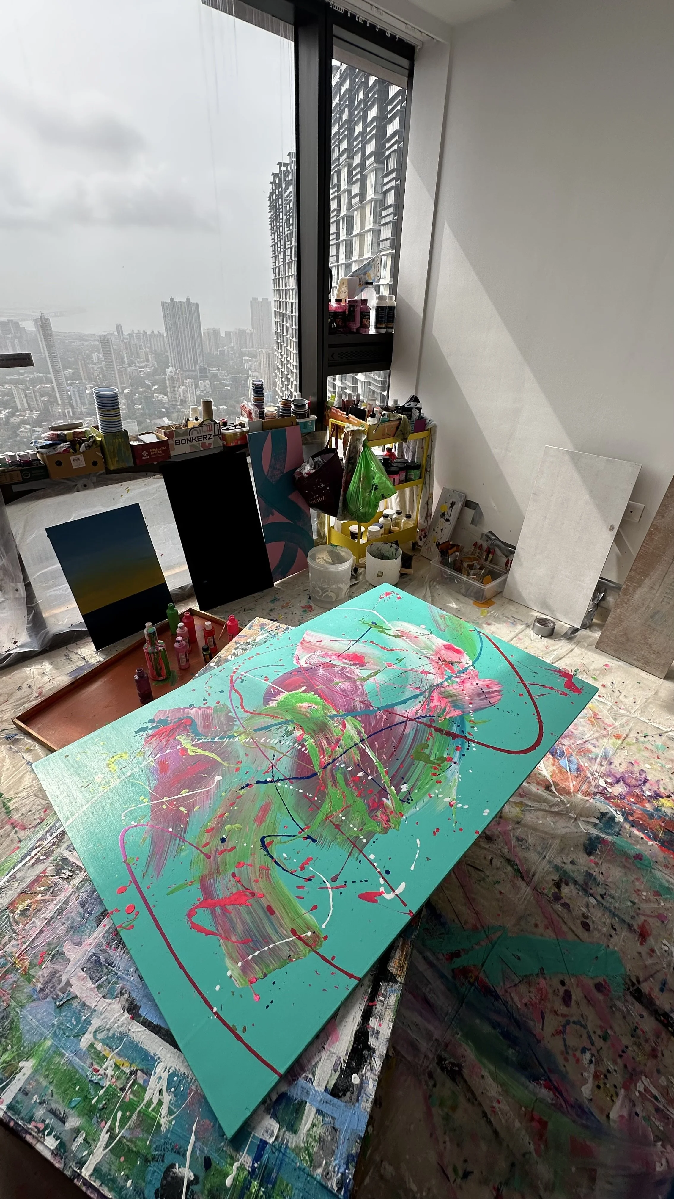 An artist's studio with a colorful abstract painting on a canvas, paint bottles on the table, and a large window overlooking a cityscape.