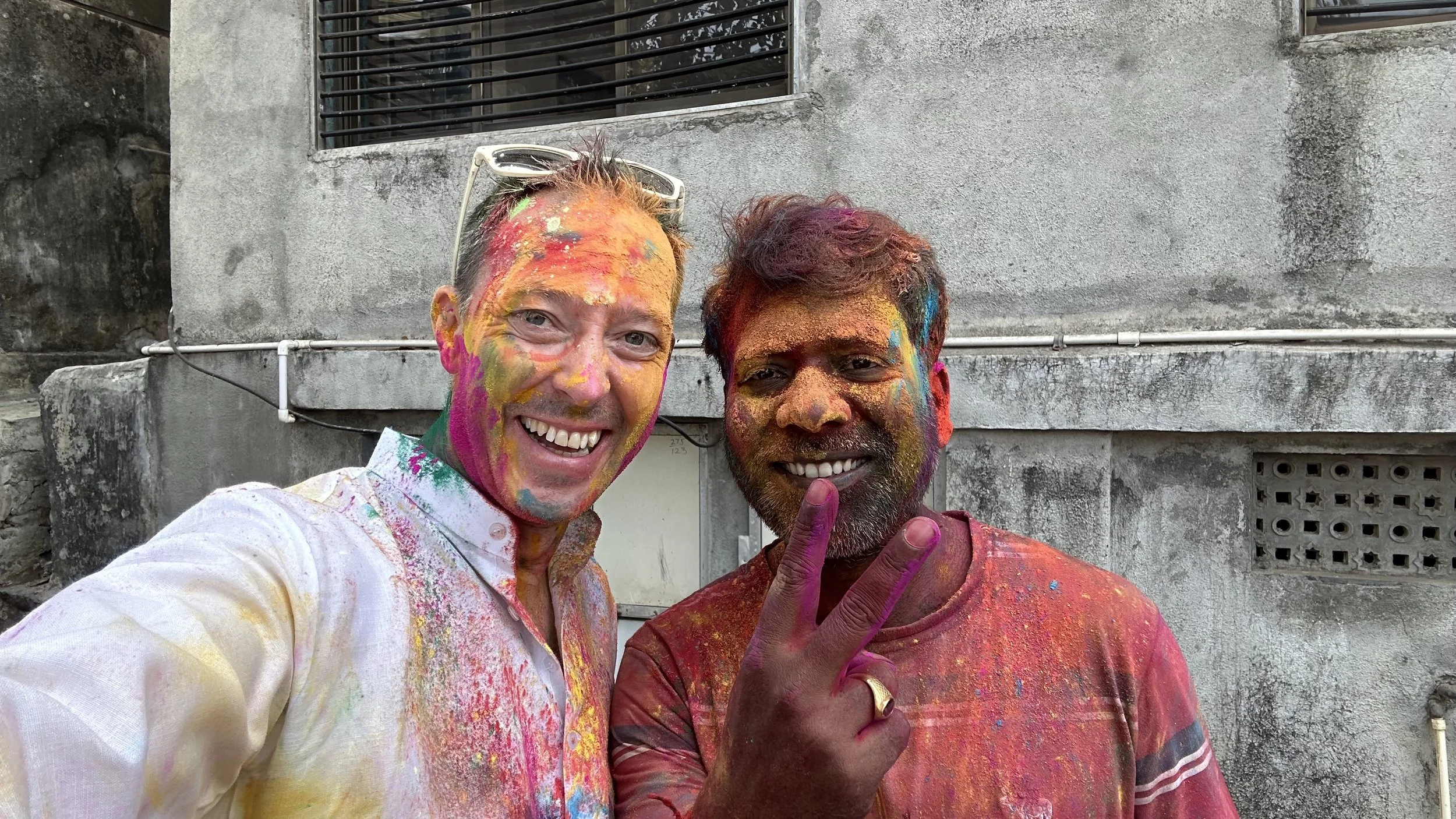 Two men smiling with colorful powders on their faces and clothes, celebrating Holi outdoors in front of a grey concrete building.
