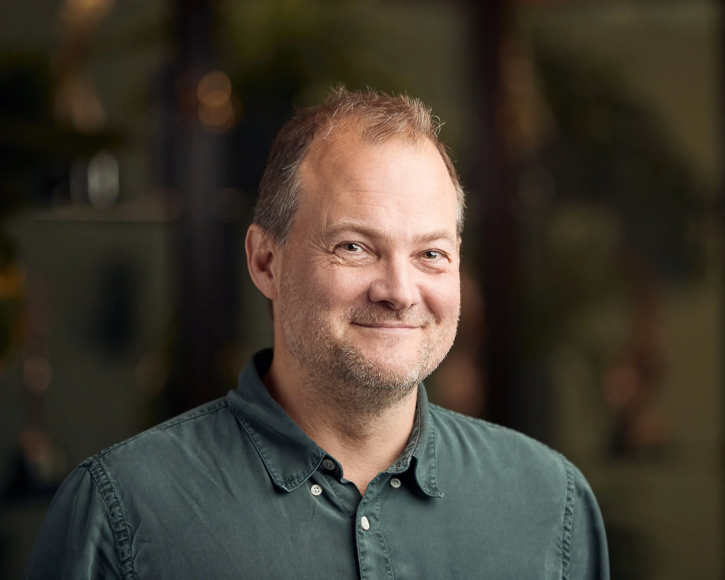 A middle-aged man with light-colored hair and a beard, wearing a button-up shirt, smiling outdoors with a blurred natural background.