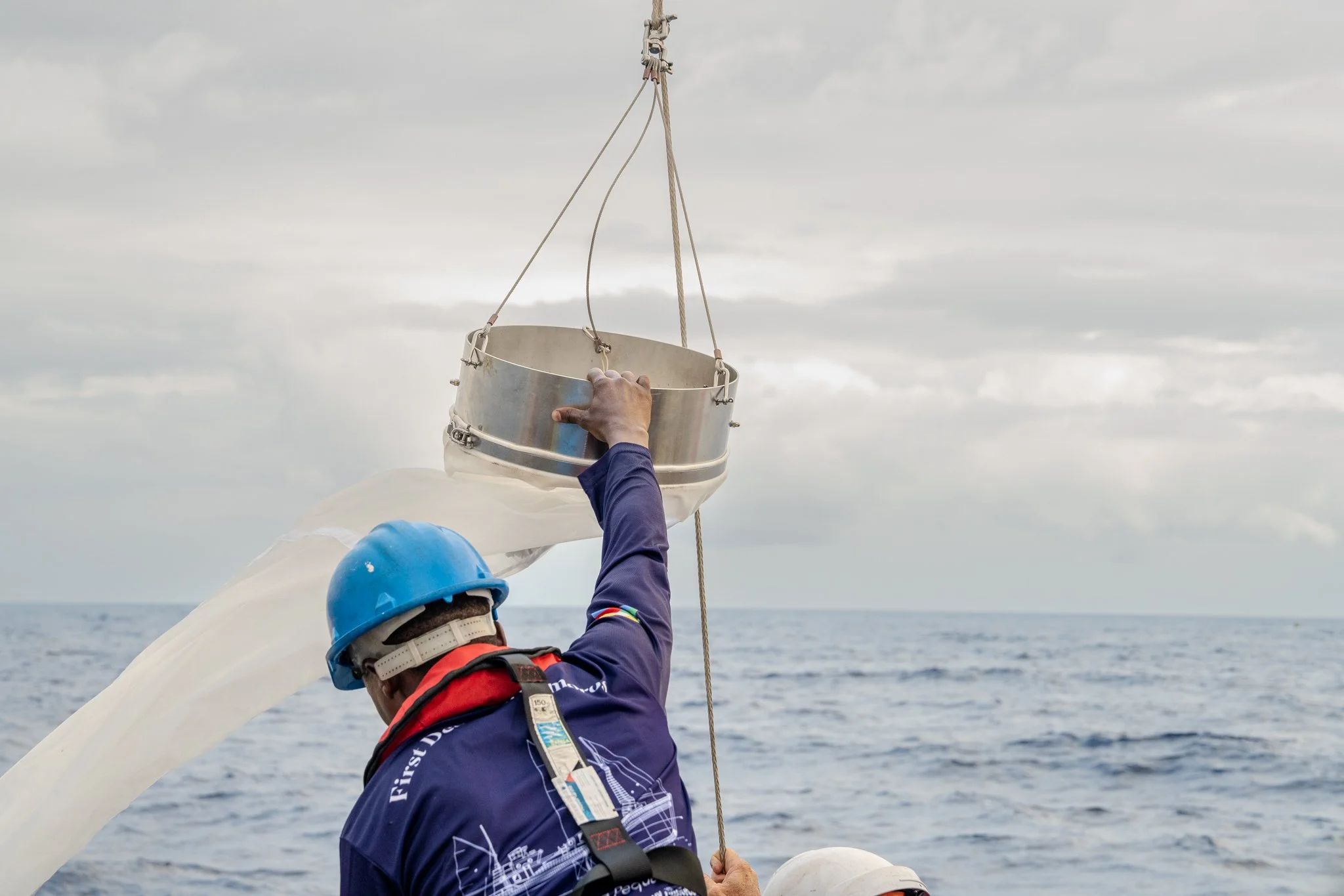 A person wearing a blue helmet and life jacket is on a boat, holding a metal bucket hanging from a rope over the water with a cloudy sky in the background.