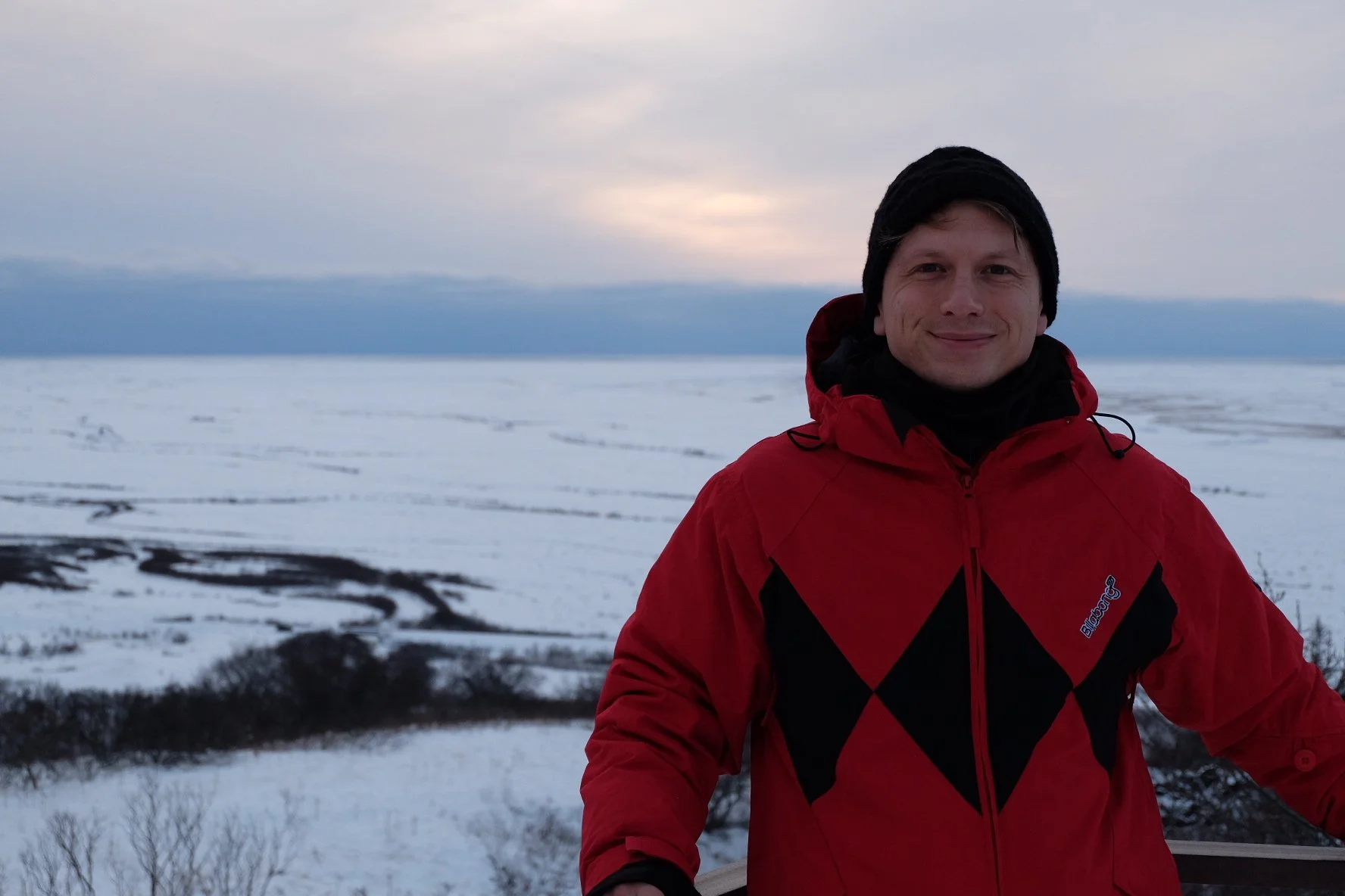 A smiling man in a red jacket and black beanie standing against a snowy landscape during sunset or sunrise, with a cloudy sky and distant snow-covered fields.