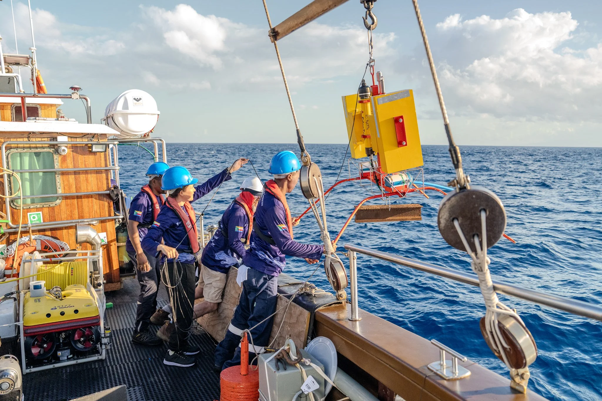 A group of five scientists in blue safety helmets working on a research vessel at sea, handling oceanographic equipment.