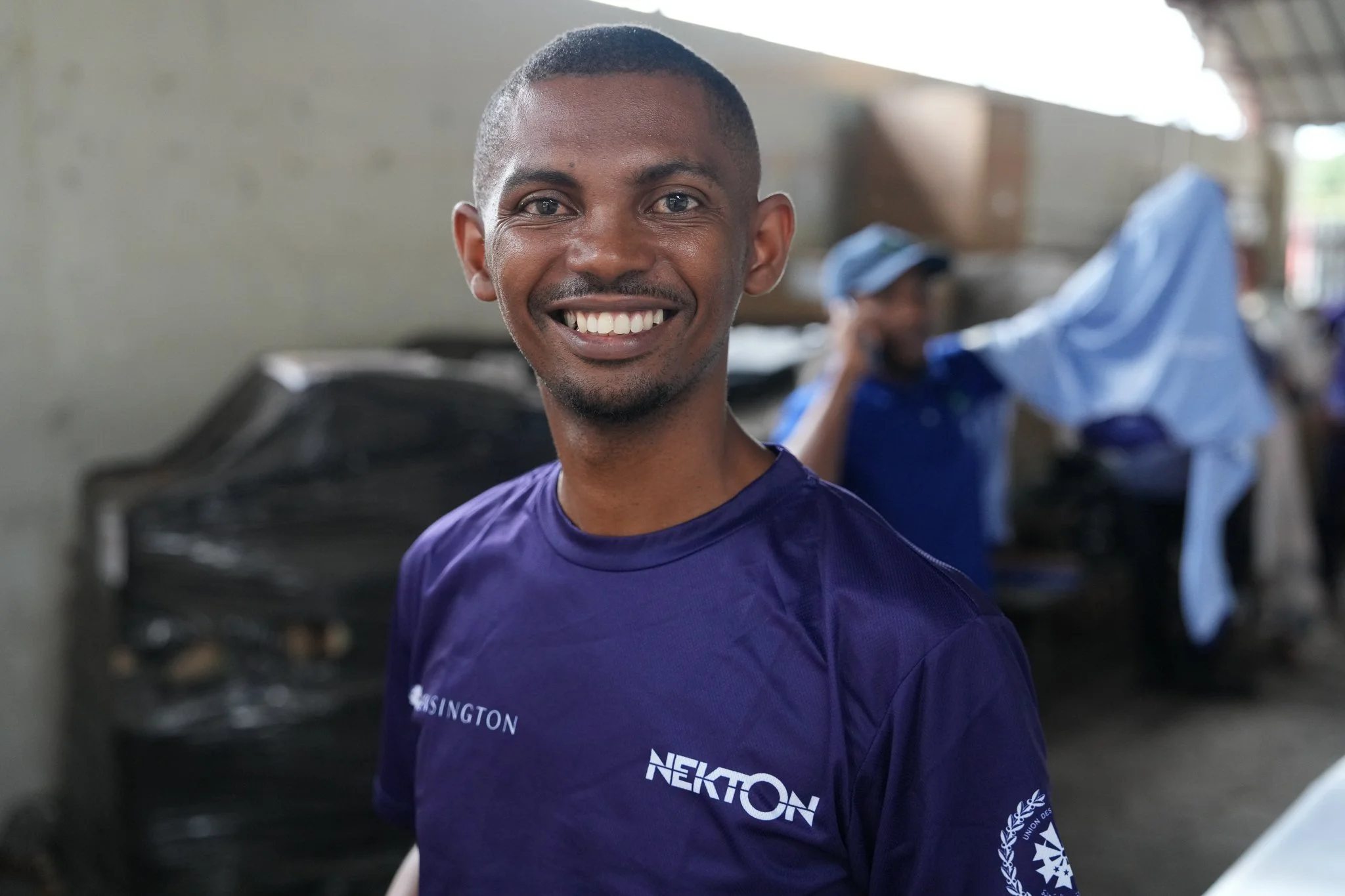 Smiling young man in a blue NEKTON sports shirt, indoors with others working in the background.