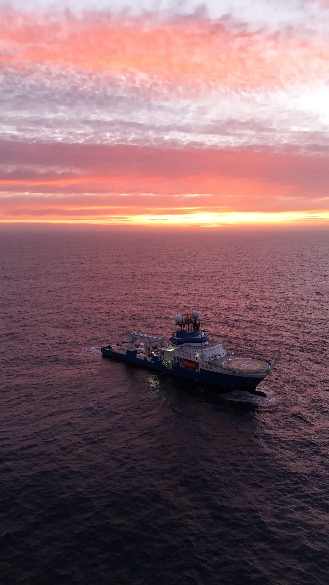 A ship with a helipad sailing on the ocean during sunset, with a colorful sky filled with pink, orange, and purple clouds.