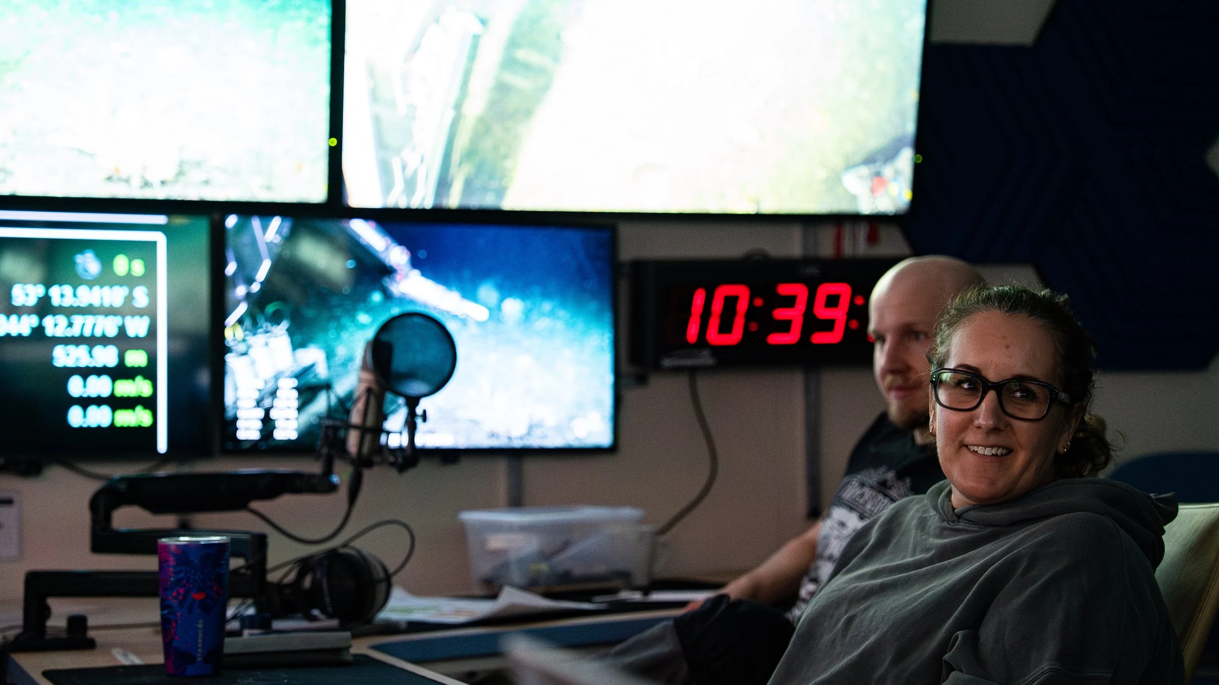 Two people sitting at a desk with monitors and a microphone, inside a control room. One woman with glasses smiling at the camera, a man next to her, and a digital clock on the wall showing 10:39.