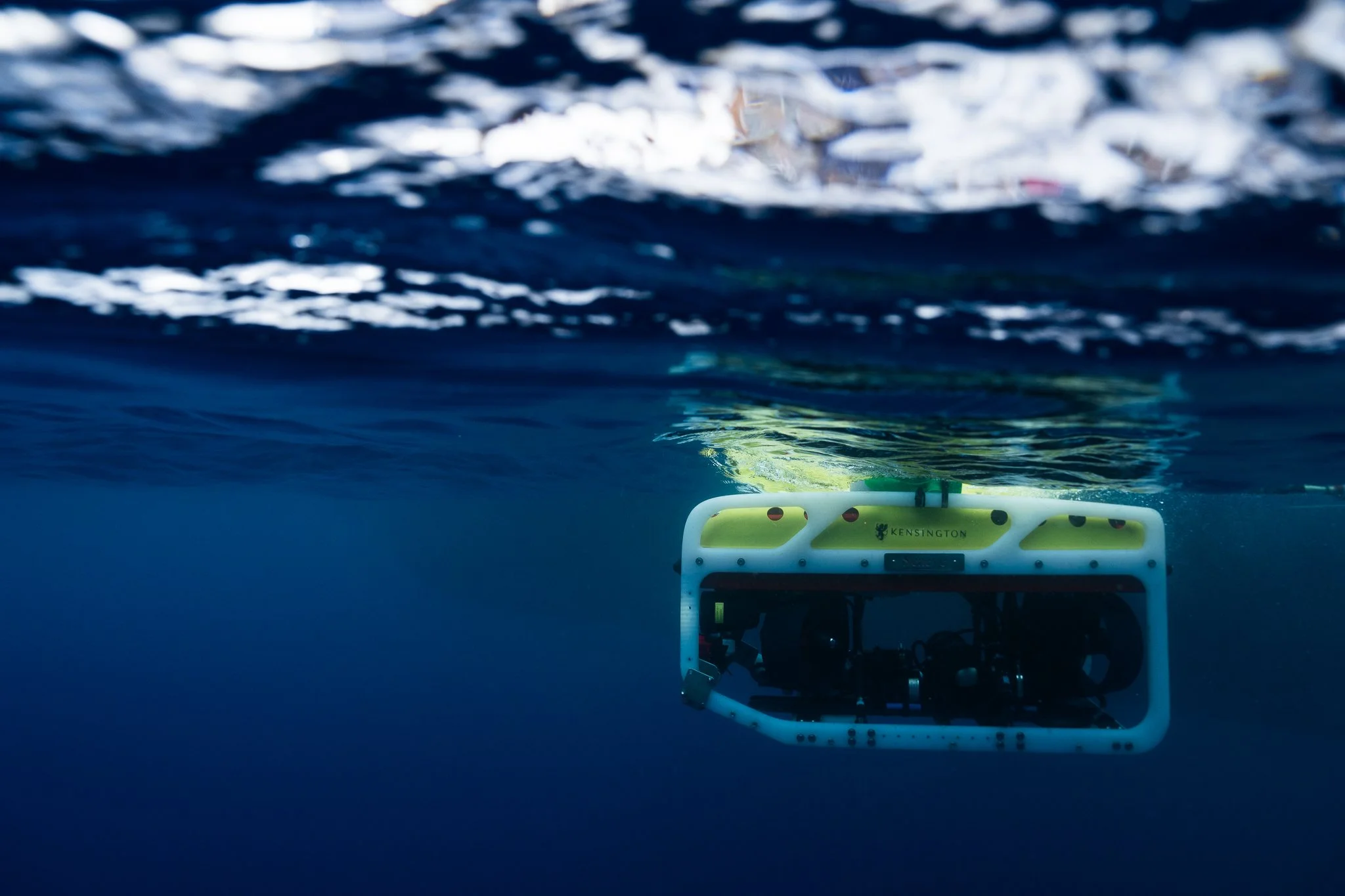 Underwater view of a ROV with a yellow top and black sensors, with the water surface above and the underwater scene below.
