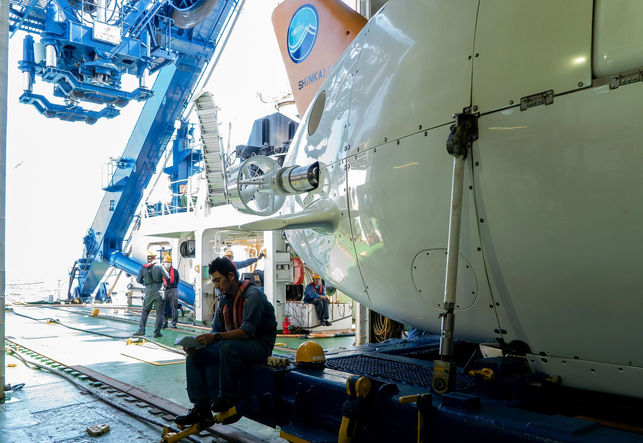 Scientists and technicians working on a large submarine or underwater vessel on a ship deck, with equipment and a blue crane in the background.