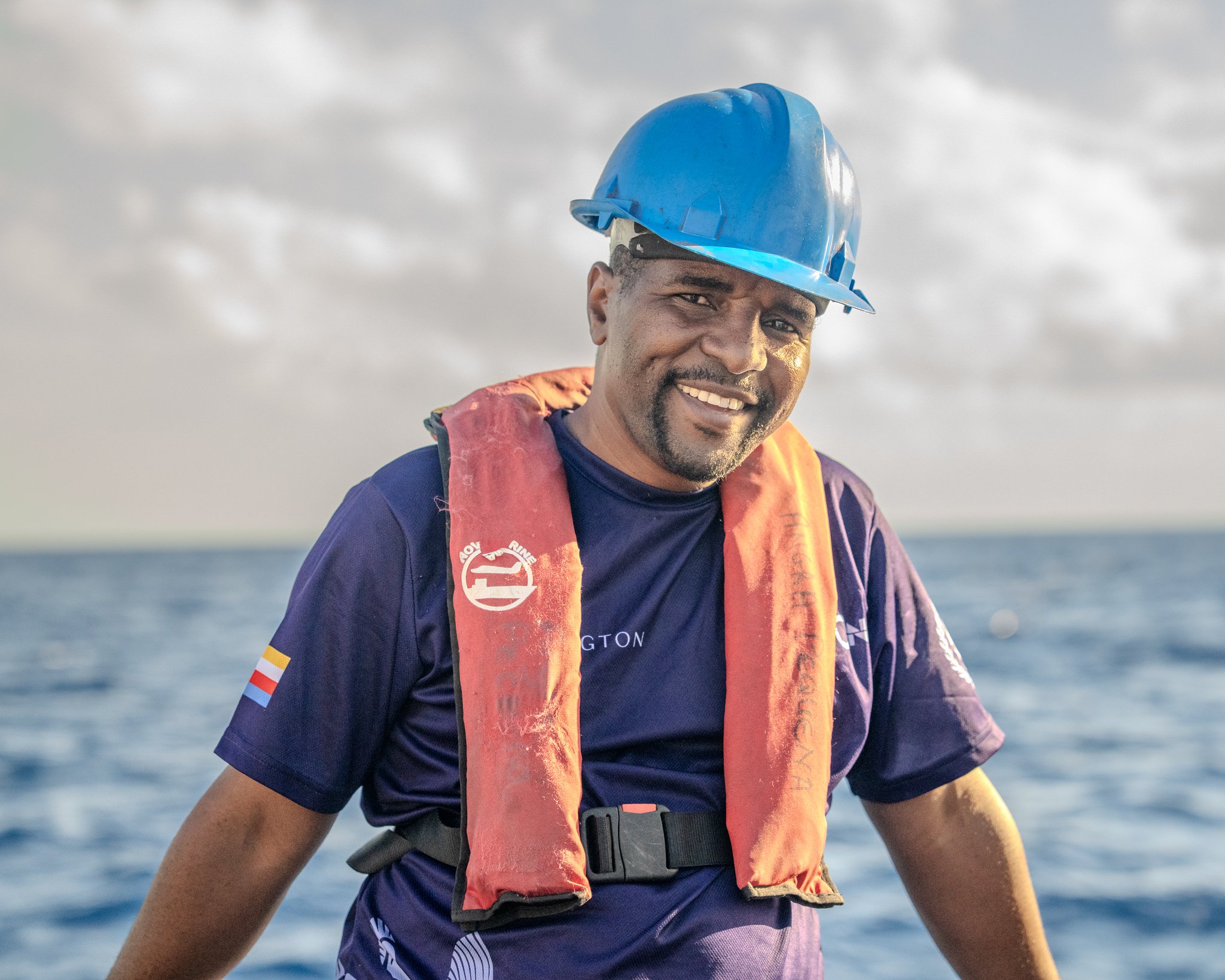 A man wearing a blue safety helmet, a navy blue shirt, and an orange life vest smiling while on a boat in the ocean.