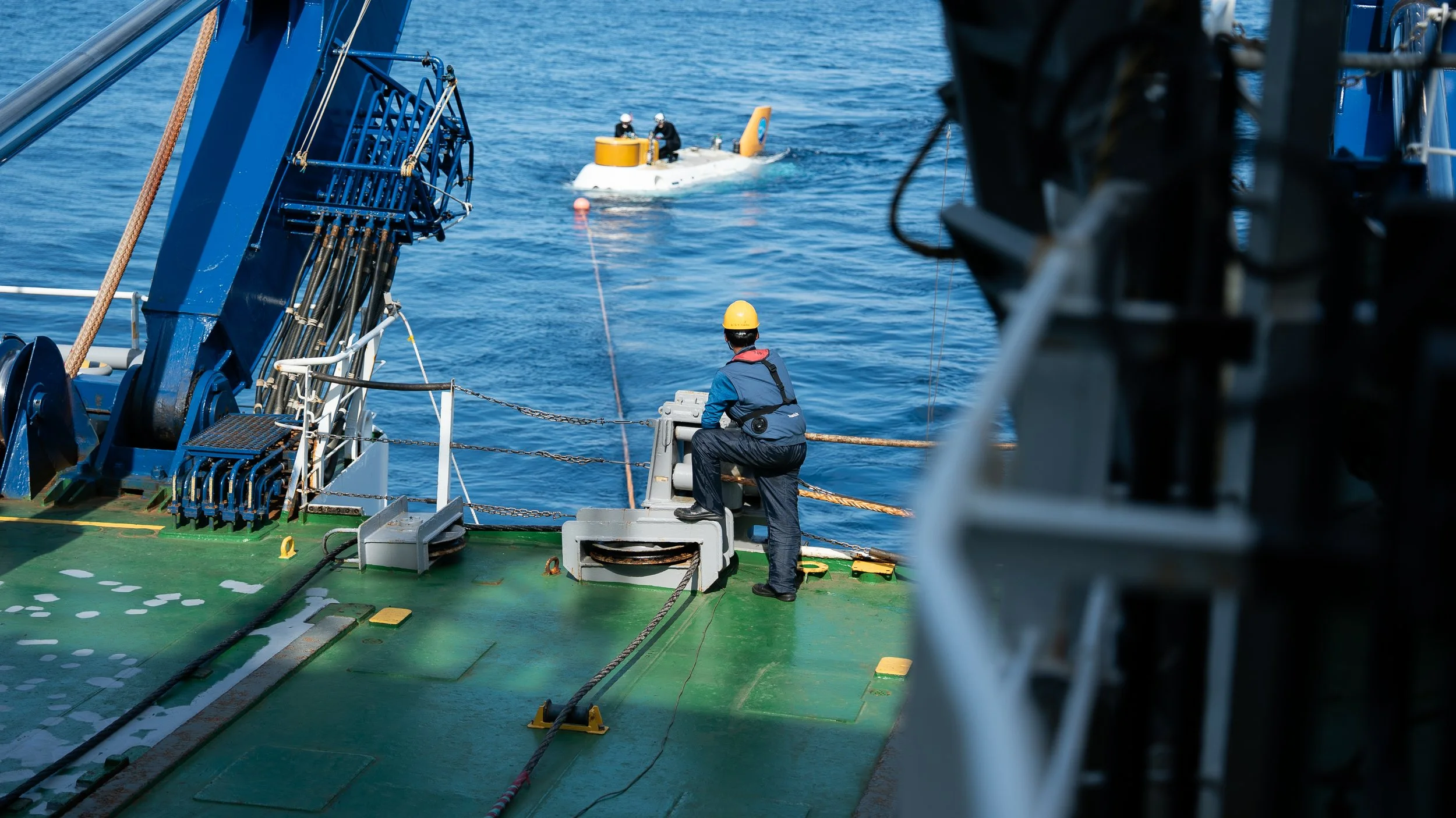 A crew member on a ship observing a submarine that is surfaced in the water, with another subgroup of people visible on the submarine, and the ocean in the background.