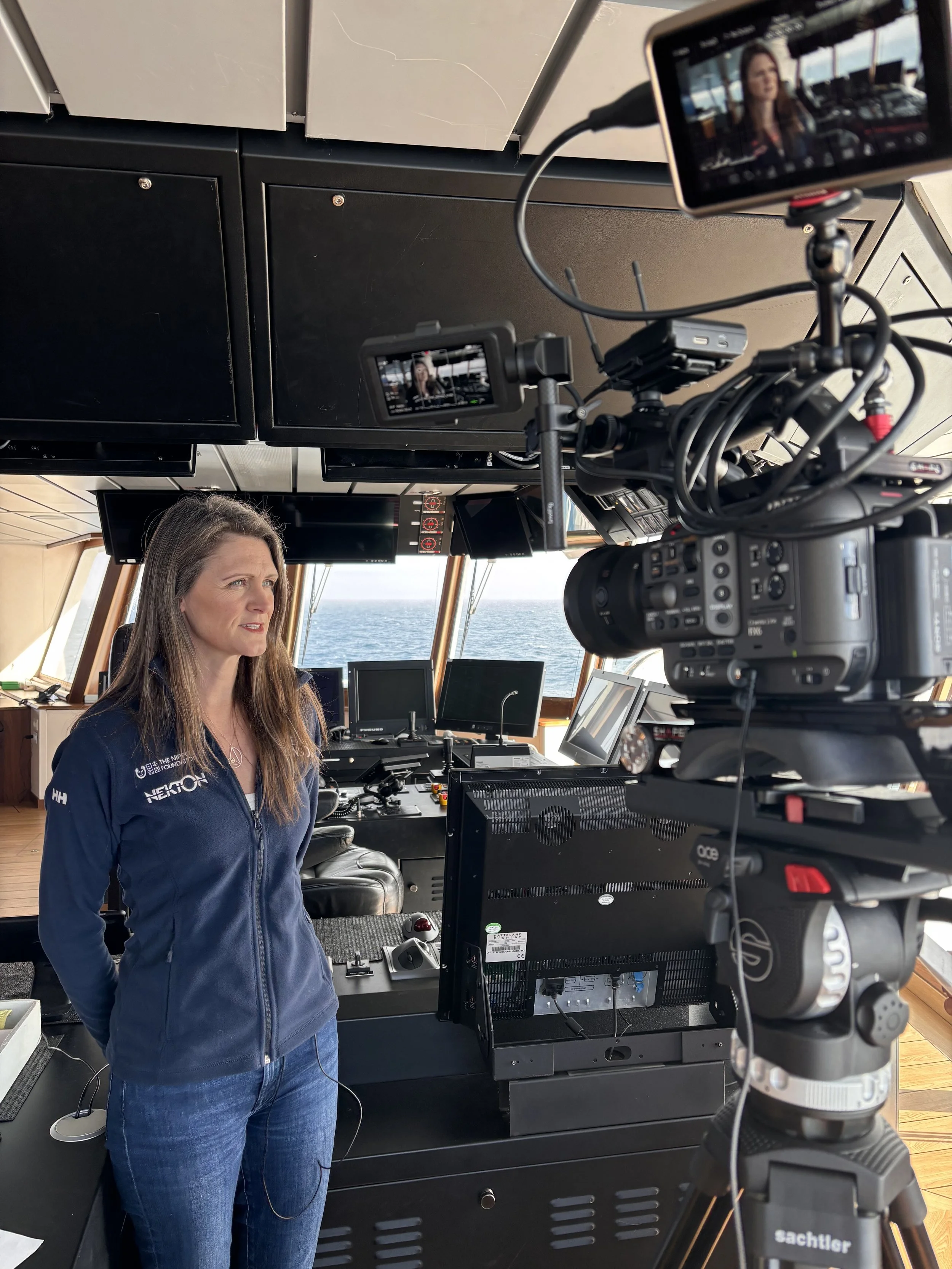 A woman in a navy blue jacket and jeans is being filmed in a ship's control room with multiple monitors and large windows showing the ocean outside.