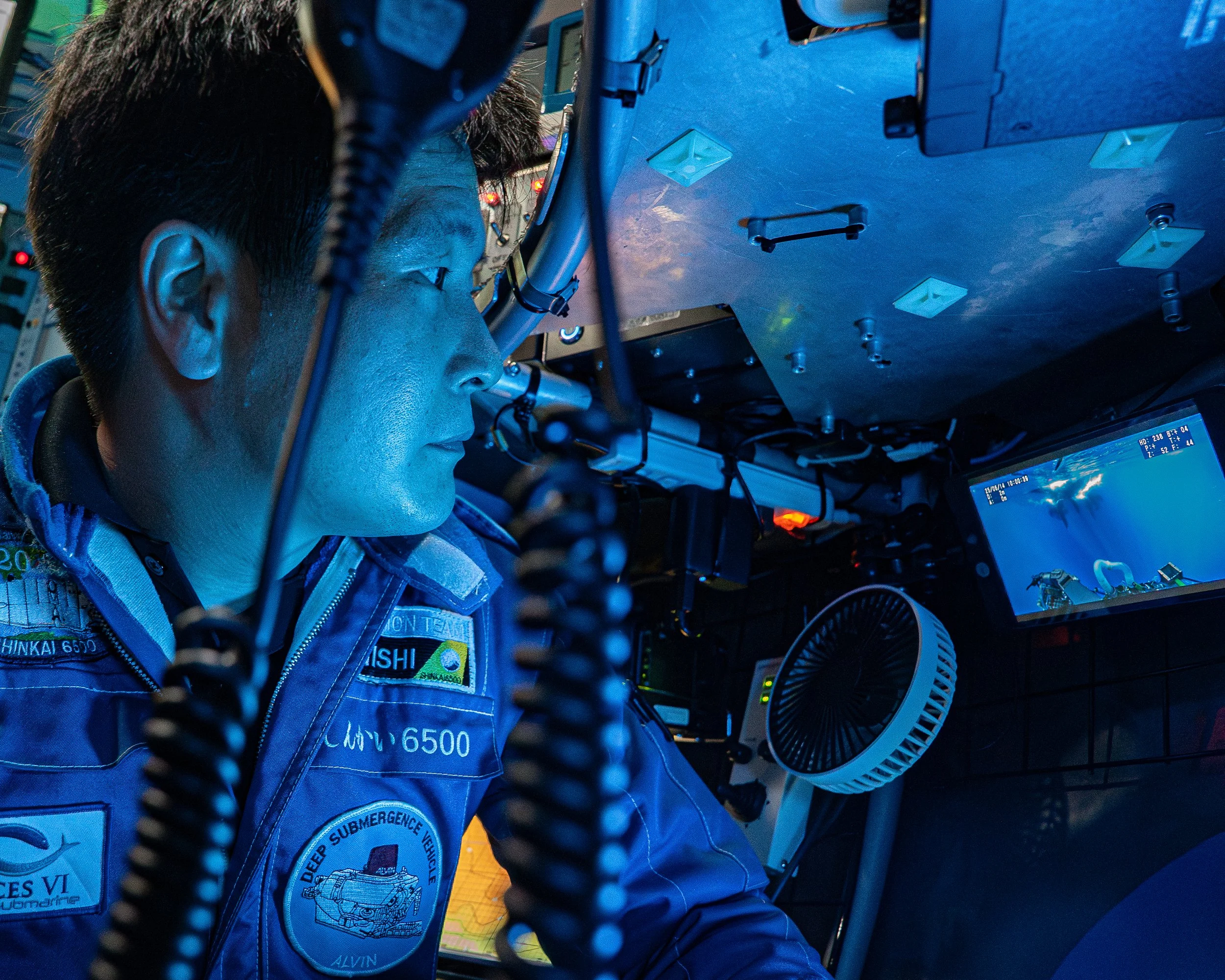 A person in a deep submergence vehicle, wearing a blue uniform, looking at screens displaying water and underwater scenes.