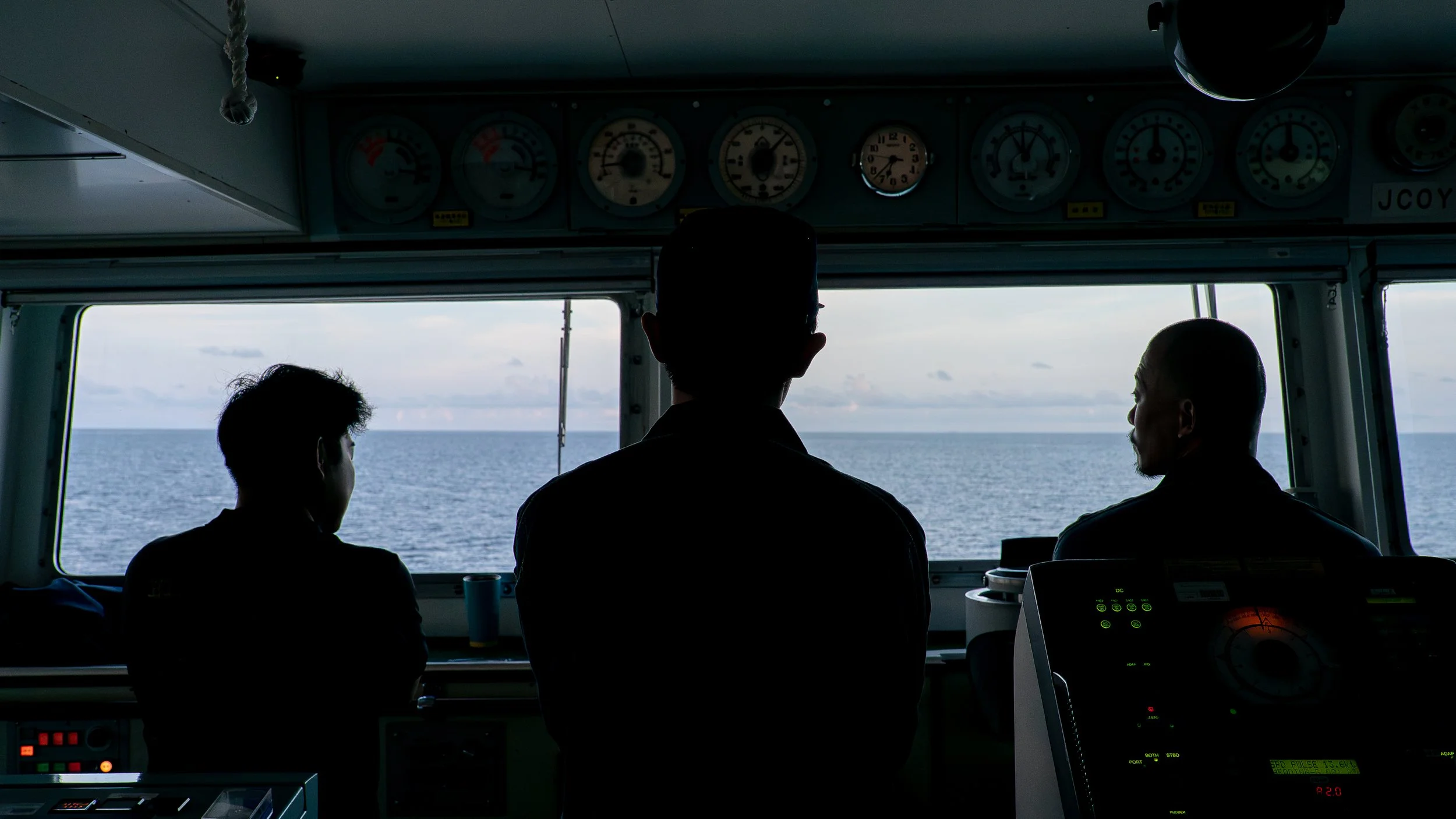 Three silhouetted individuals in a ship's control room looking out at the ocean through large front windows, with various gauges and controls in the cockpit.