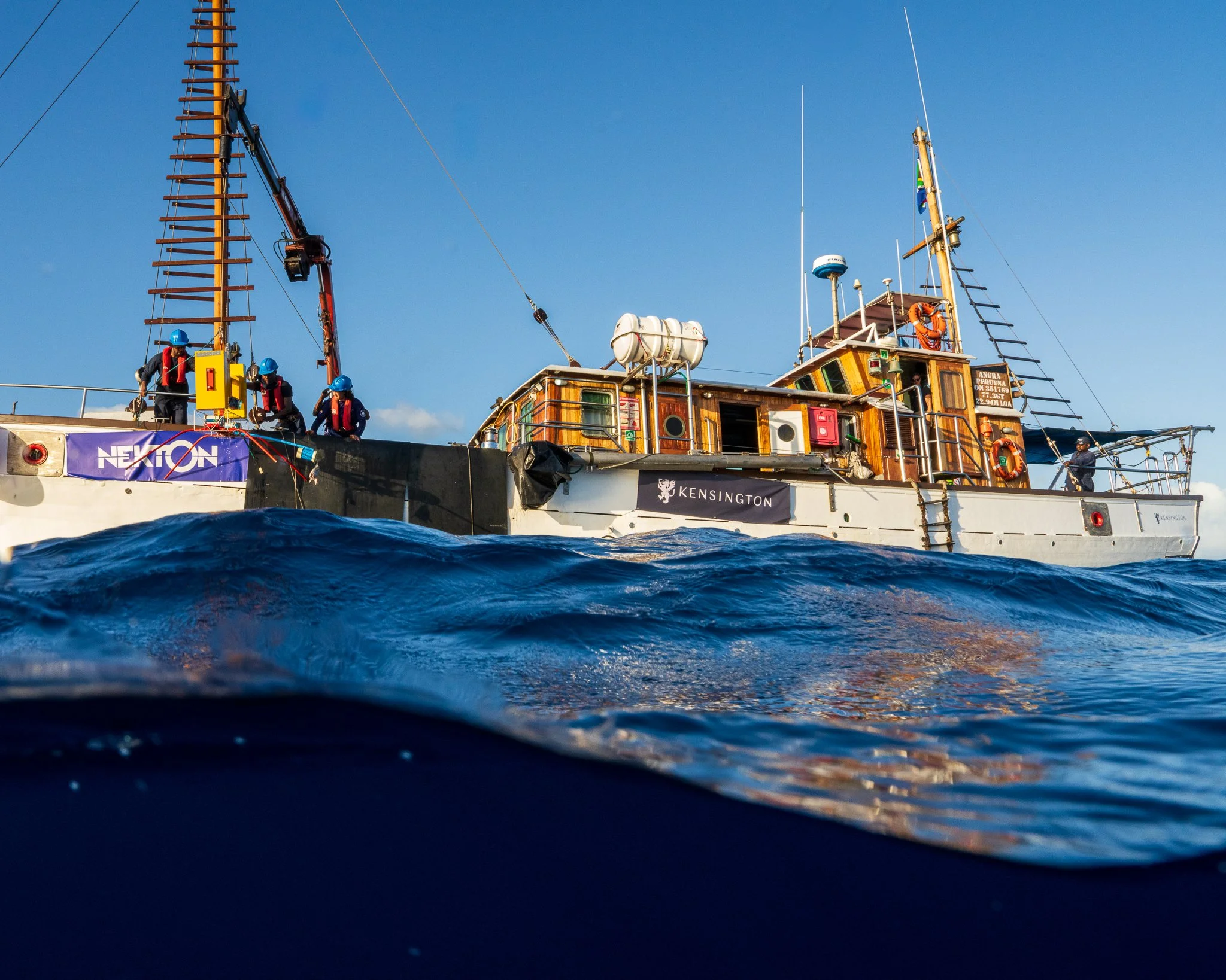 A boat with Kensington and Nekton branding, with several crew members wearing safety gear, floating on the ocean with waves, under a clear blue sky.