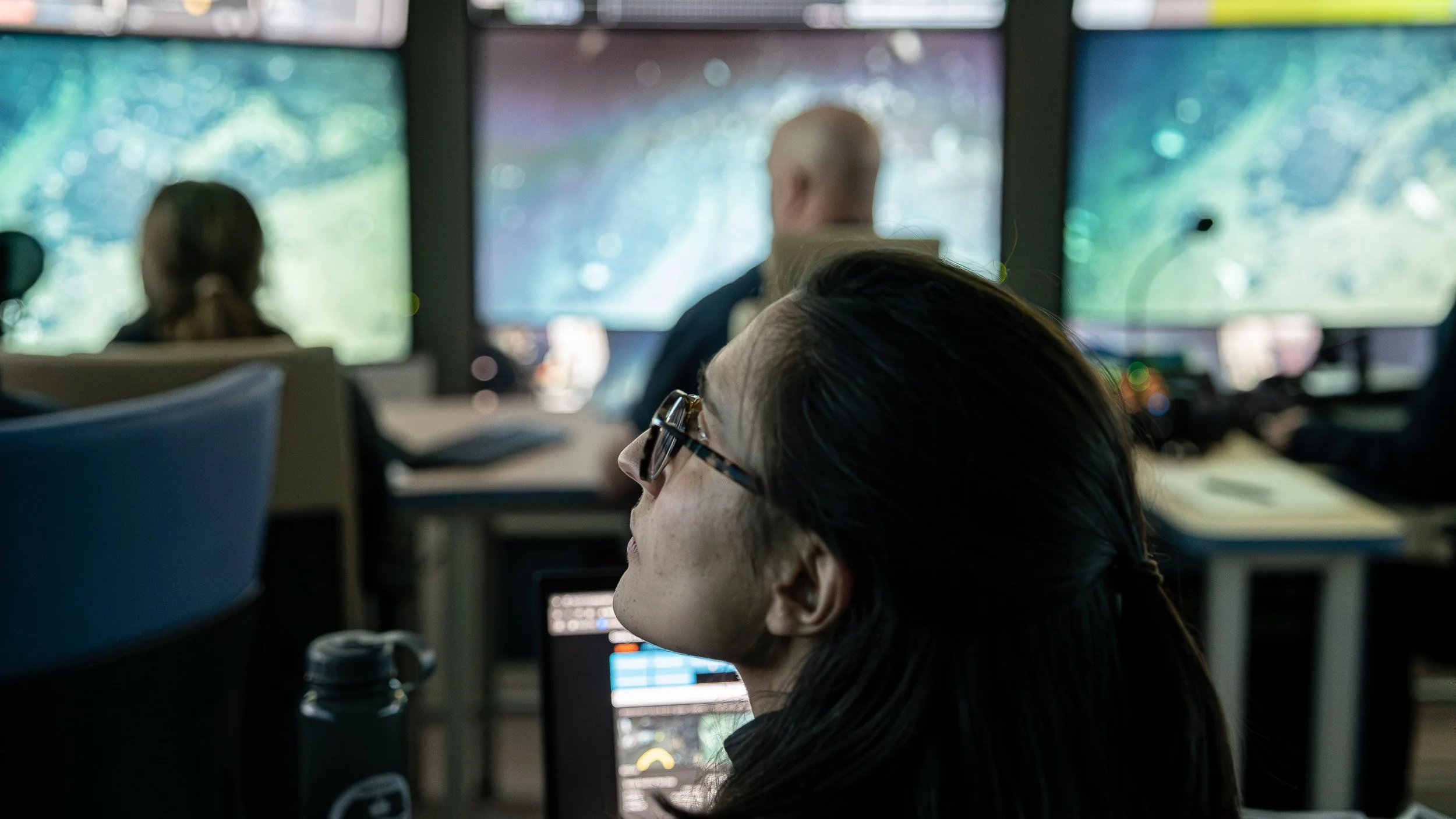 A woman with dark hair and glasses working at a desk in an office, with multiple large monitors displaying complex graphical data or software interfaces.