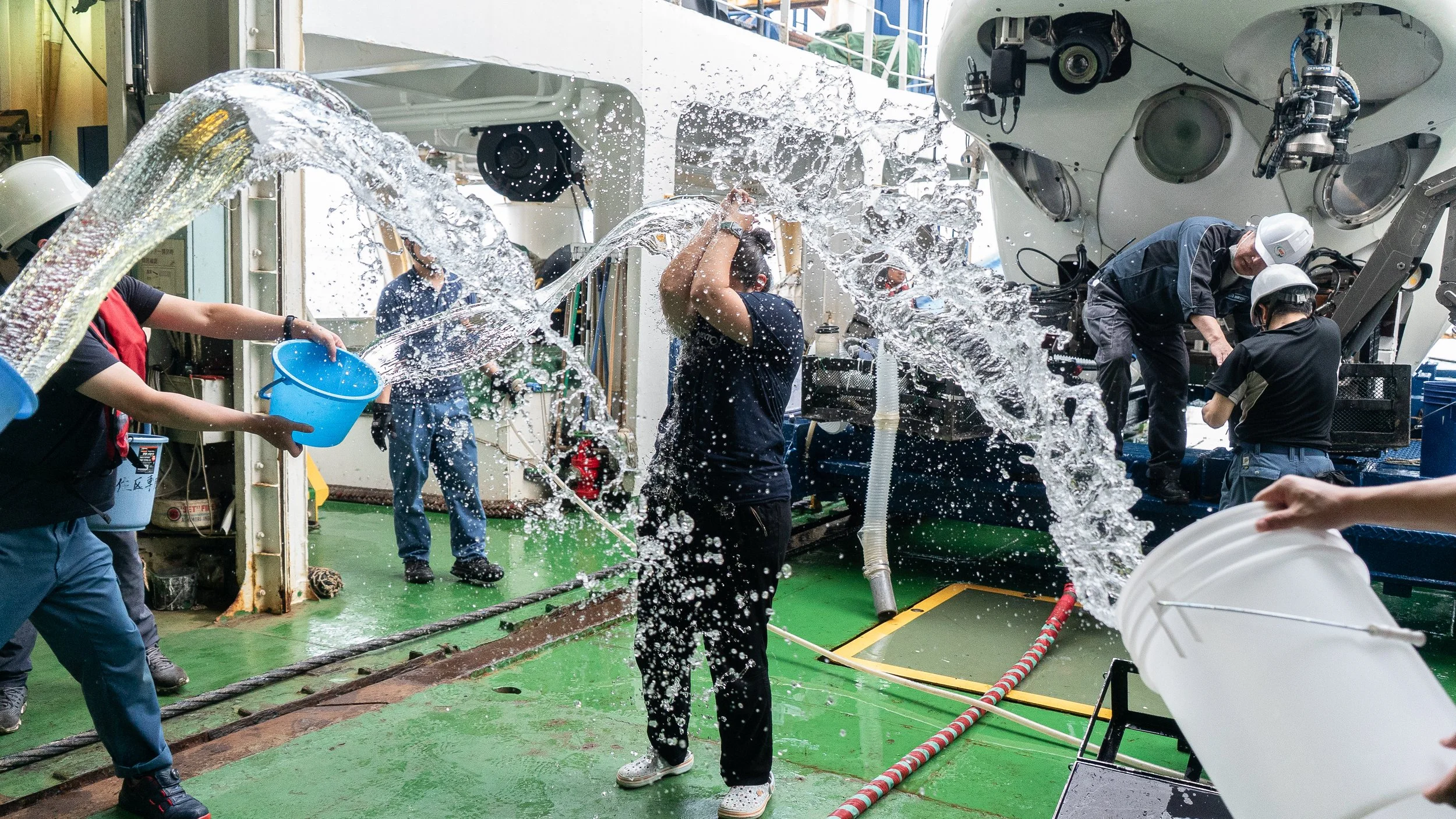 People working on a boat, with water being splashed around in a busy industrial environment, some wearing safety helmets.