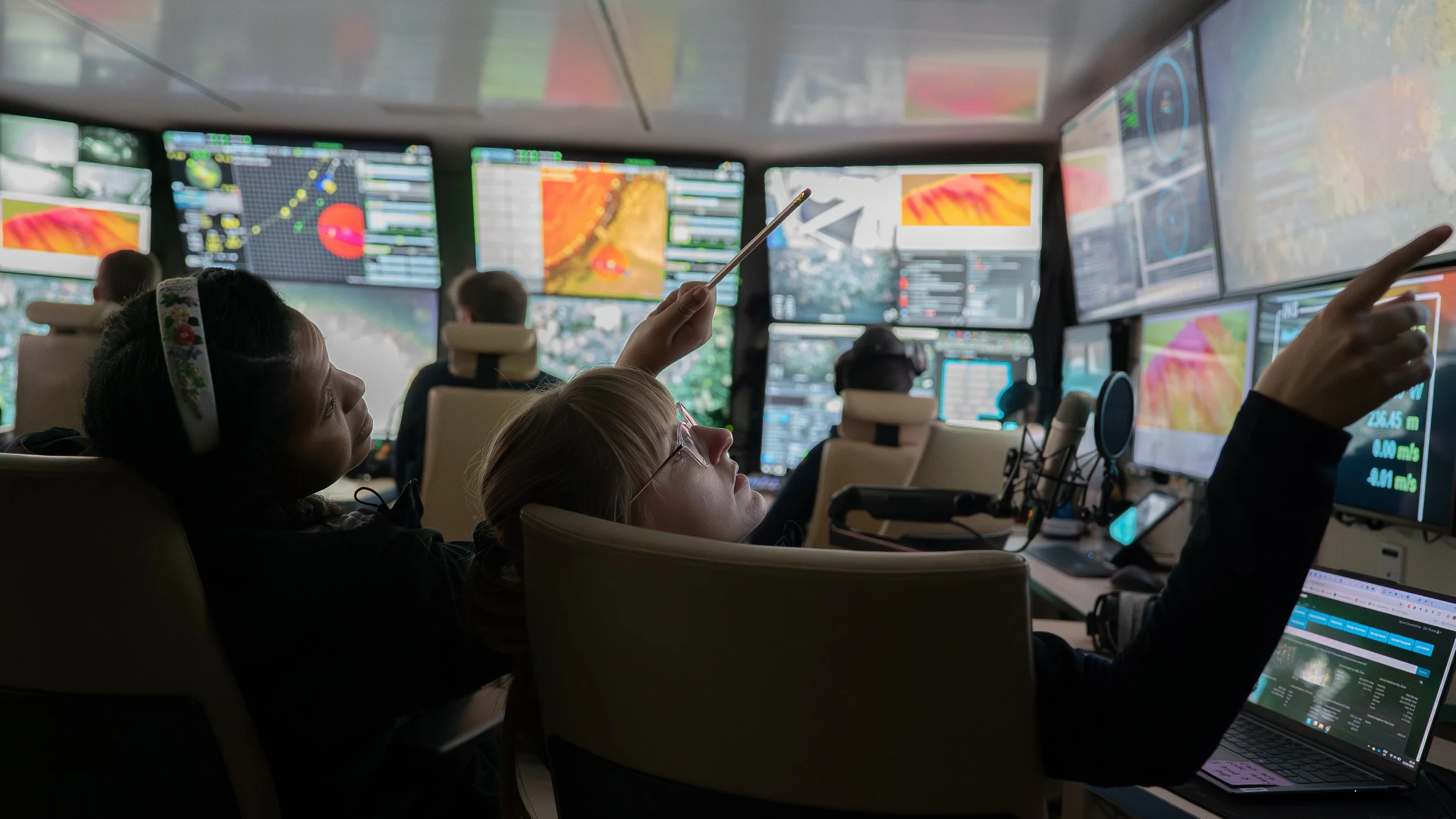 Control room with multiple monitors displaying data, graphics, and maps. Several people are seated, working with screens and equipment.
