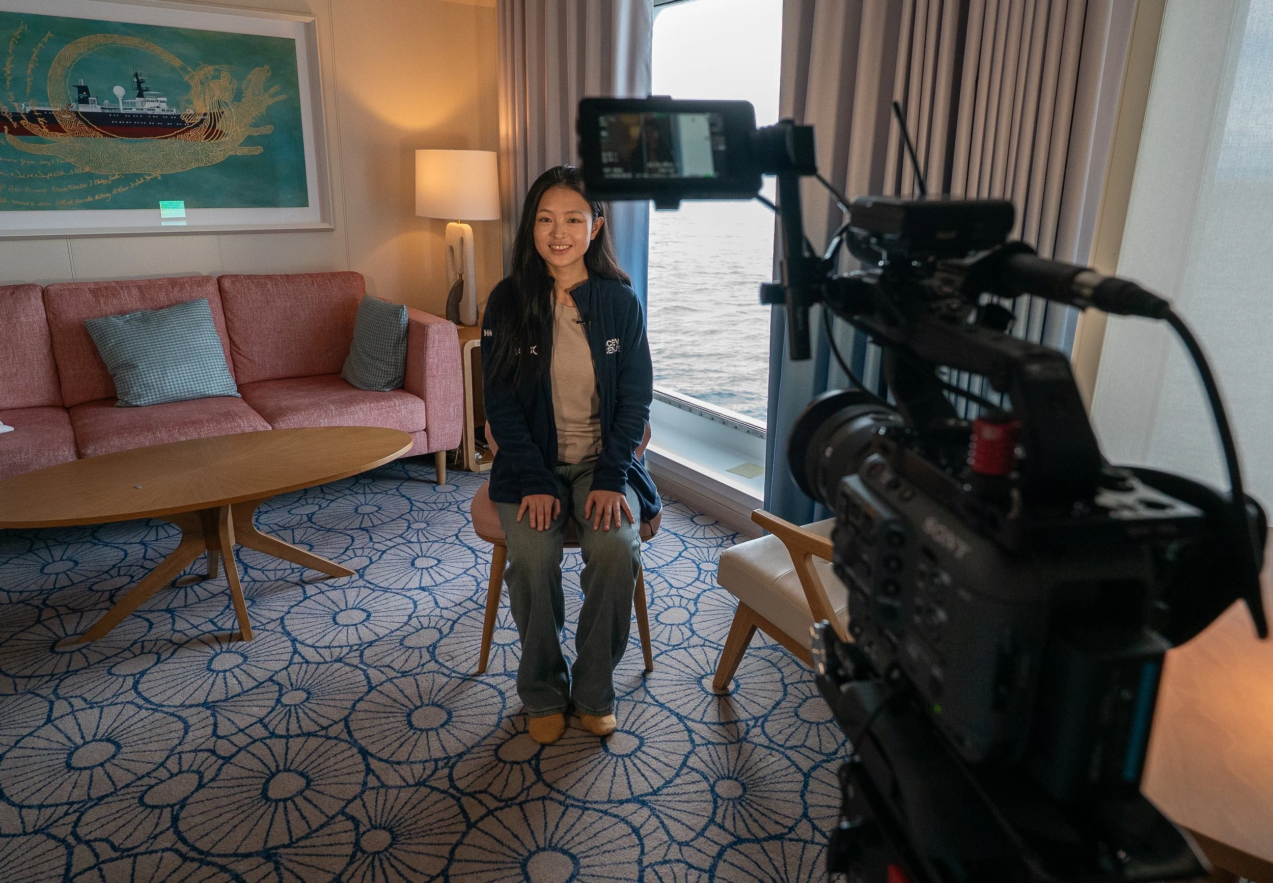 Woman sitting on a chair in a room with a large window showing a body of water, a pink sofa with pillows, a coffee table, a patterned carpet, a standing lamp, and artwork on the wall, while a camera captures her interview.