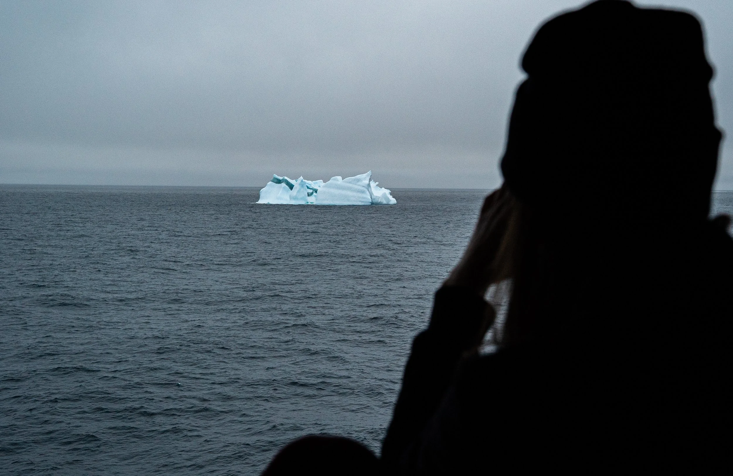 Person in silhouette looking at an iceberg floating in the ocean on a cloudy day.