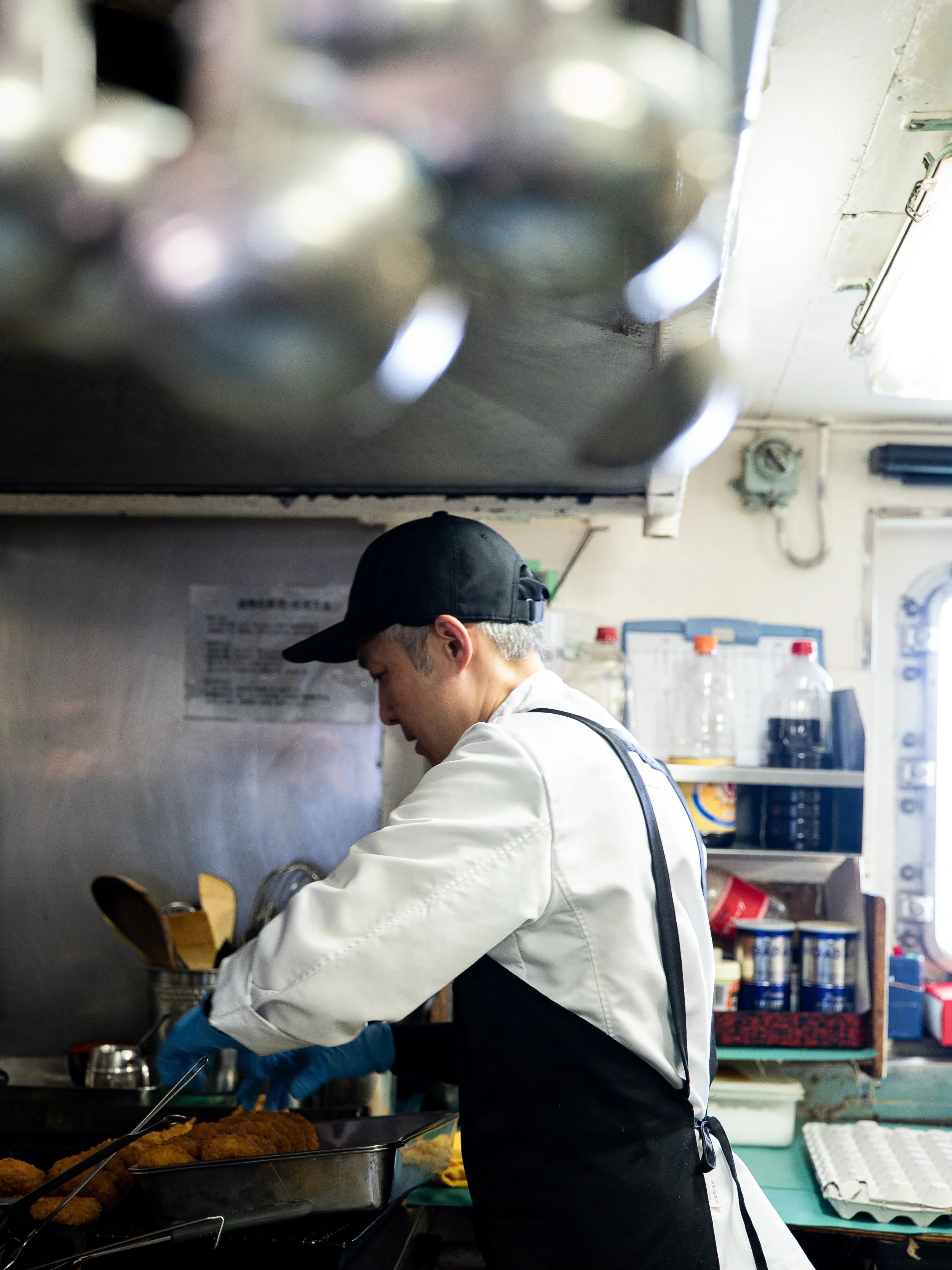 A chef wearing a black cap, white shirt, black apron, and blue gloves prepares fried food in a kitchen.