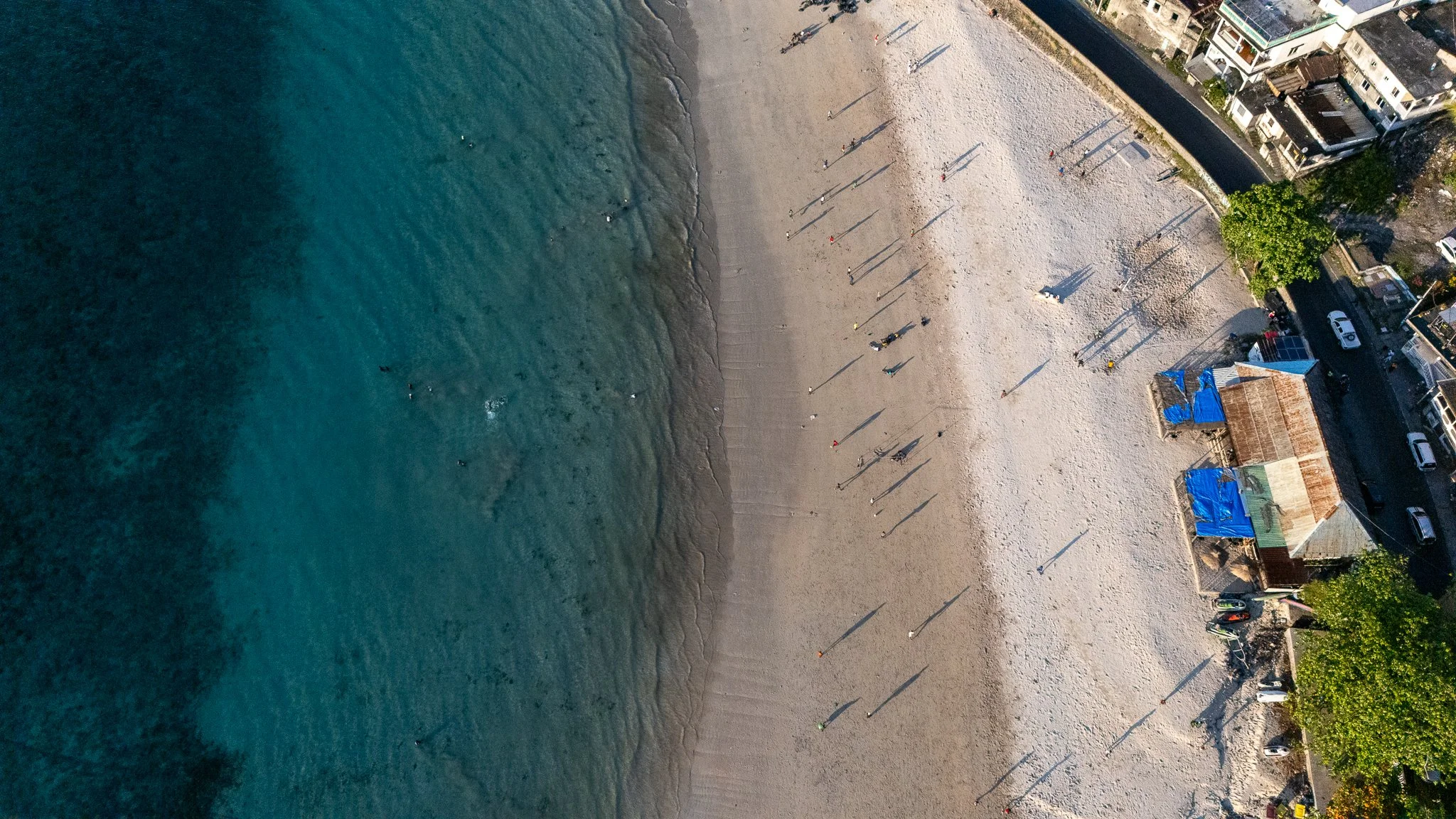 Aerial view of a beach with people walking and swimming near the shore, adjacent to a row of buildings and parked cars.