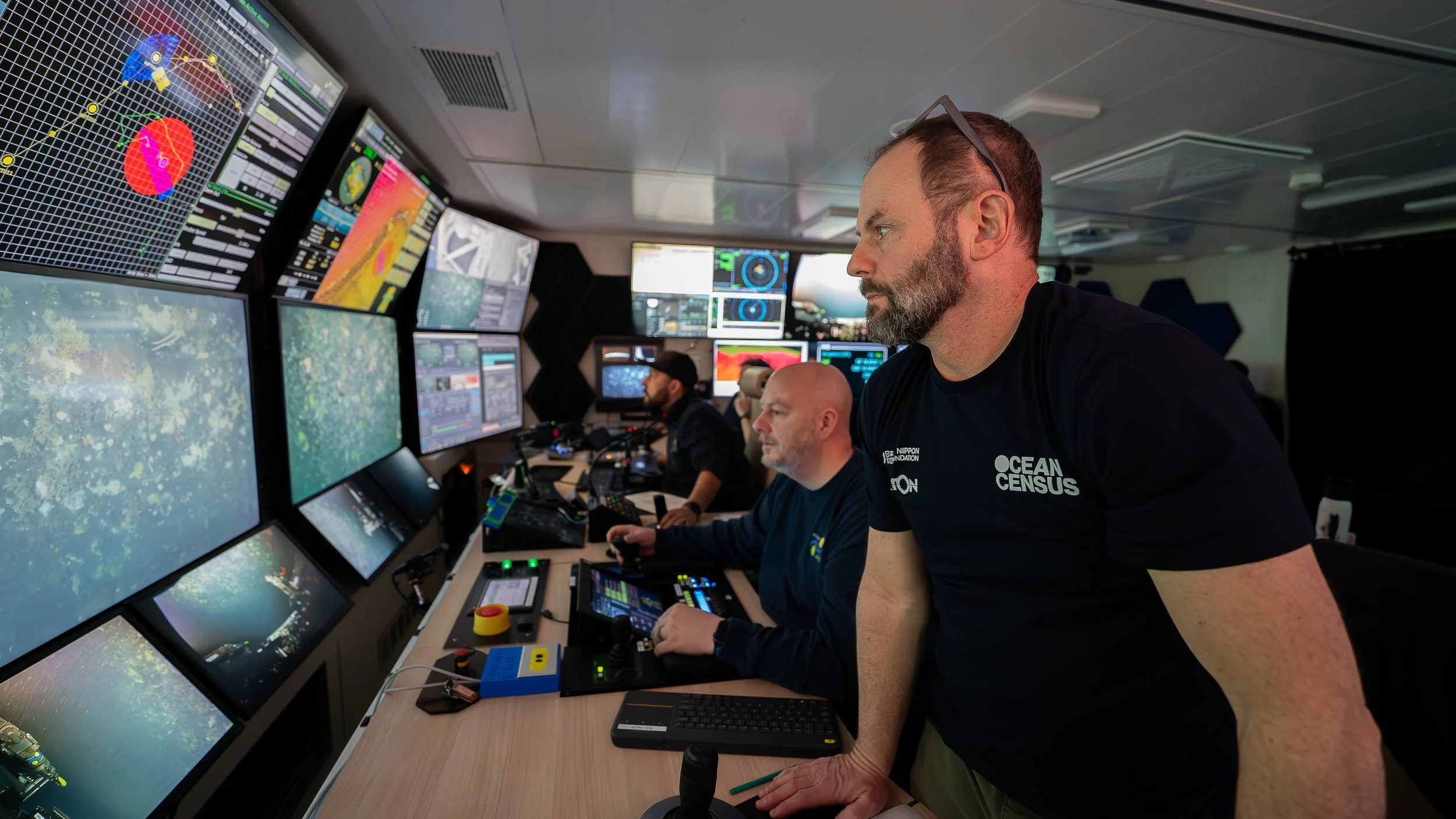 Three men working at a multi-monitor control room with various data and graphics displays, one in the foreground wearing a black Ocean Census t-shirt, the others in the background operating equipment.