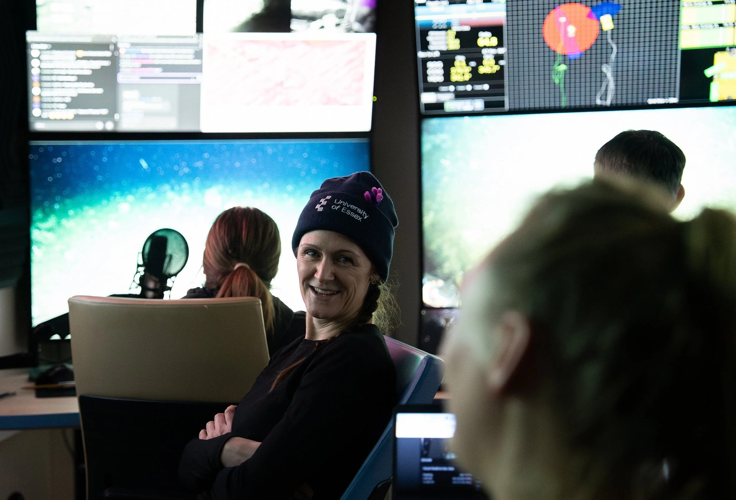 A woman smiling and wearing a University of Essex beanie sitting in a control room with multiple monitors displaying various data and graphs.
