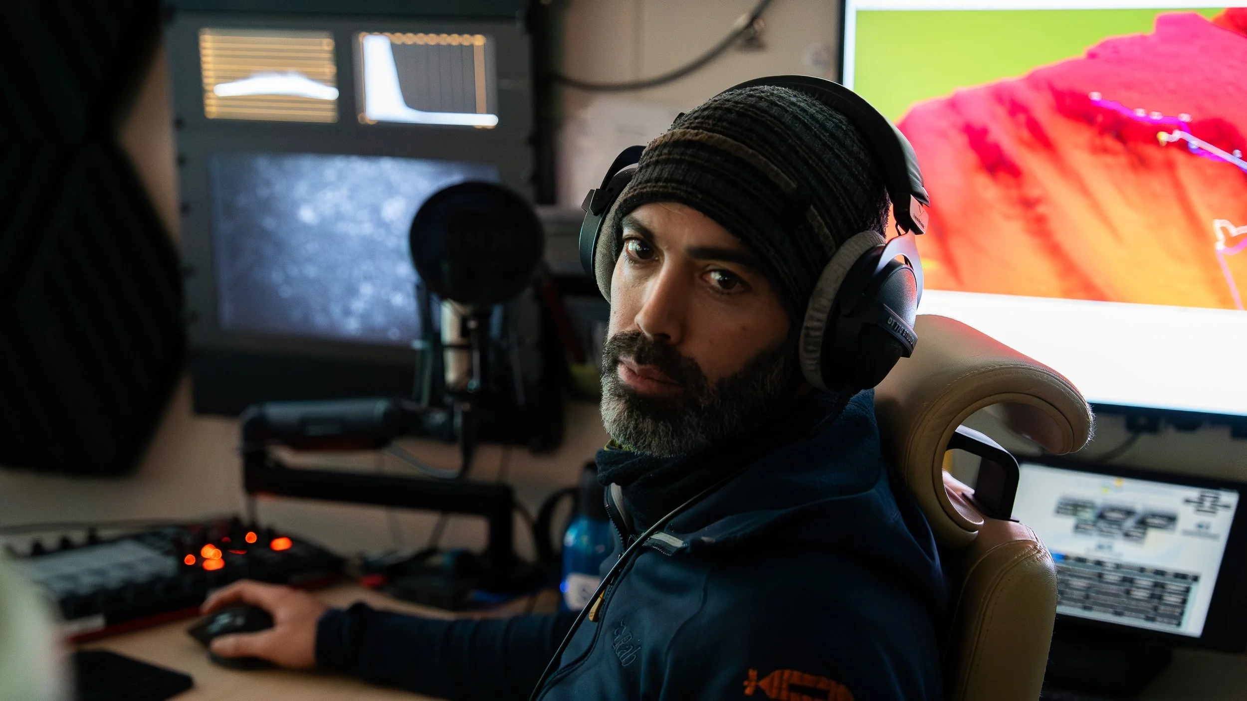 Man with dark beard wearing headphones, black beanie, and dark jacket working at a computer station with multiple monitors and audio equipment in a dimly lit room.