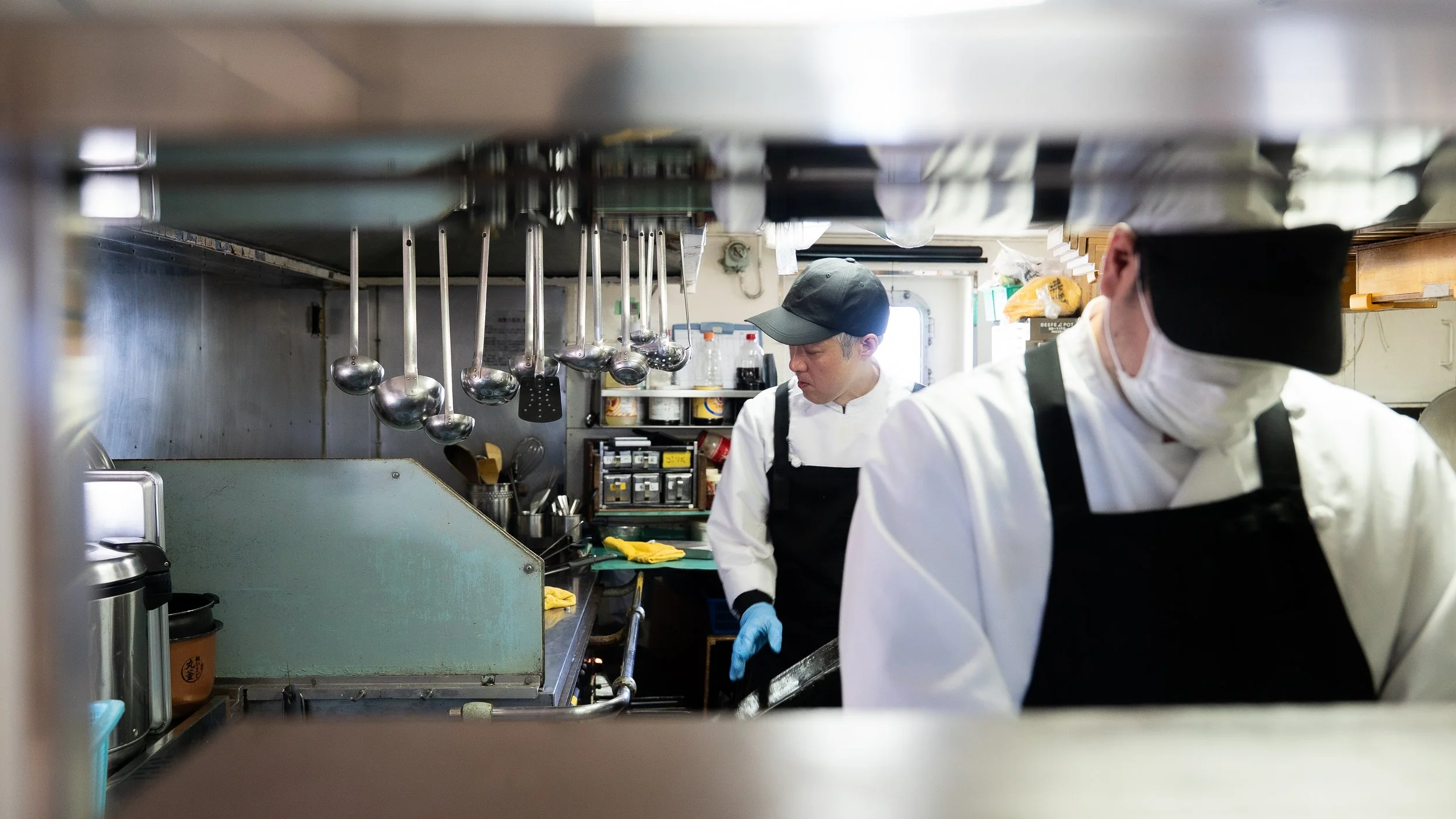 Two chefs wearing white uniforms, black aprons, and hats working in a busy kitchen, seen through a framing of kitchen equipment and utensils.