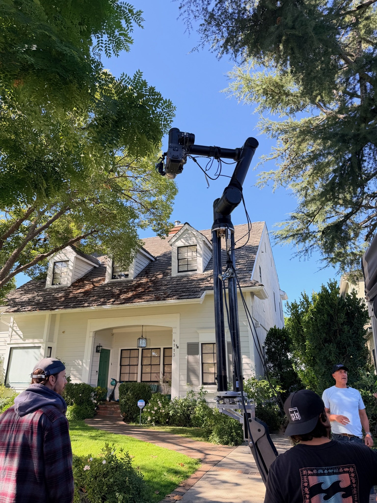 Film or camera crew setting up equipment in a residential neighborhood, with a towering camera crane pointing towards a white house with dormer windows, surrounded by trees and greenery.