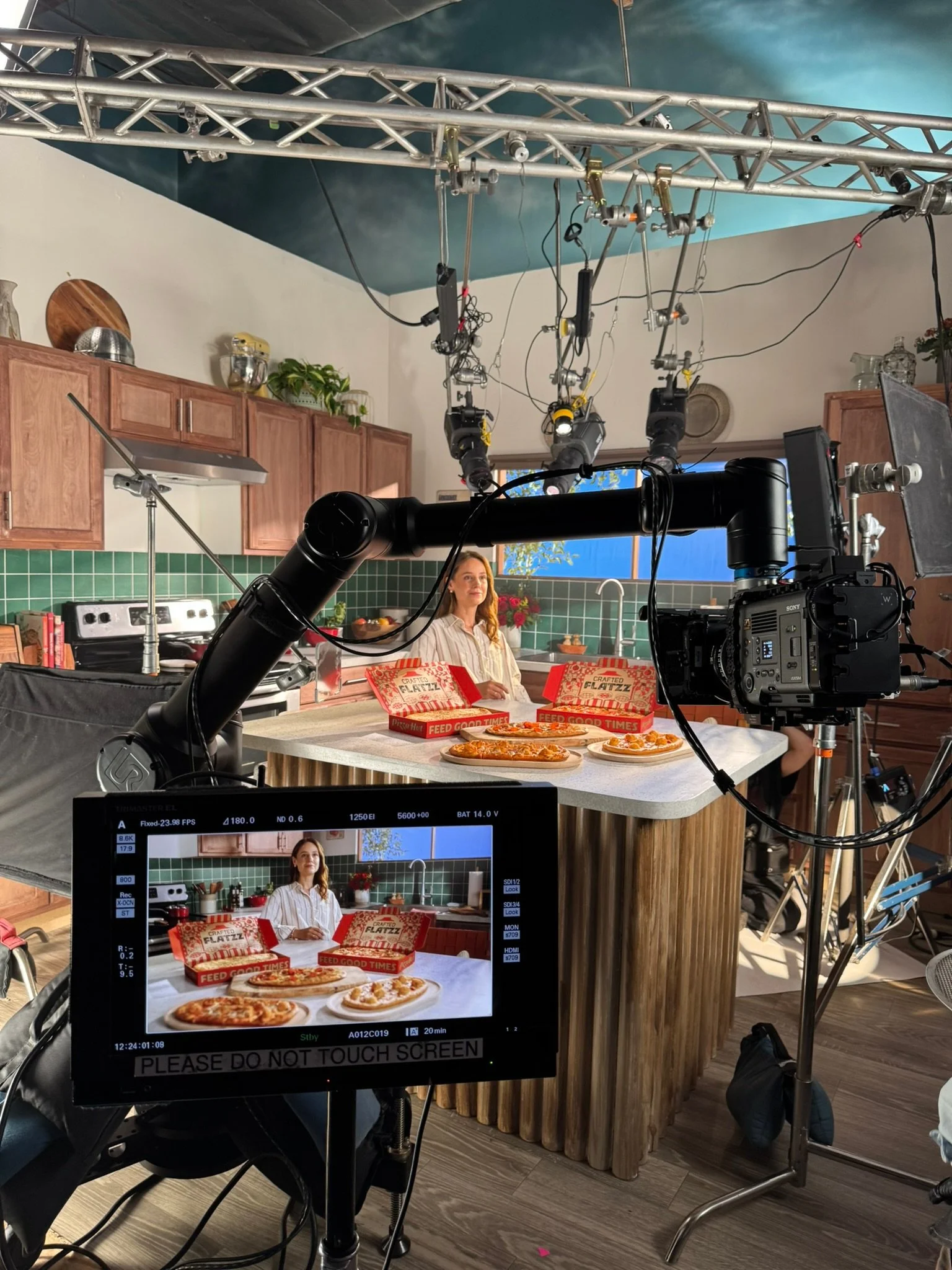 Behind the scenes of a cooking show with a woman sitting at a kitchen island with pizzas and pizza boxes on it, captured on a monitor and camera setup.