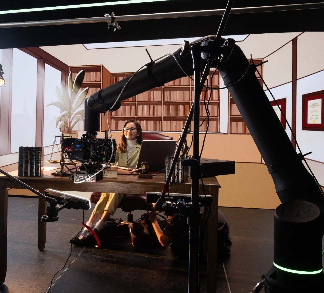 A woman sits at a desk on a film set, smiling, with a background depicting an office with bookshelves and framed certificates, and a large robotic arm and filming equipment in the foreground.