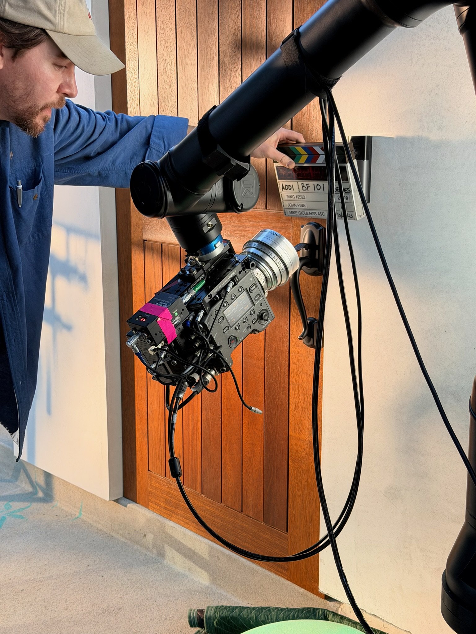 A filmmaker adjusting settings on a professional camera mounted on a road rig with a matte box, positioned against a wall with wood paneling and a film slate nearby.