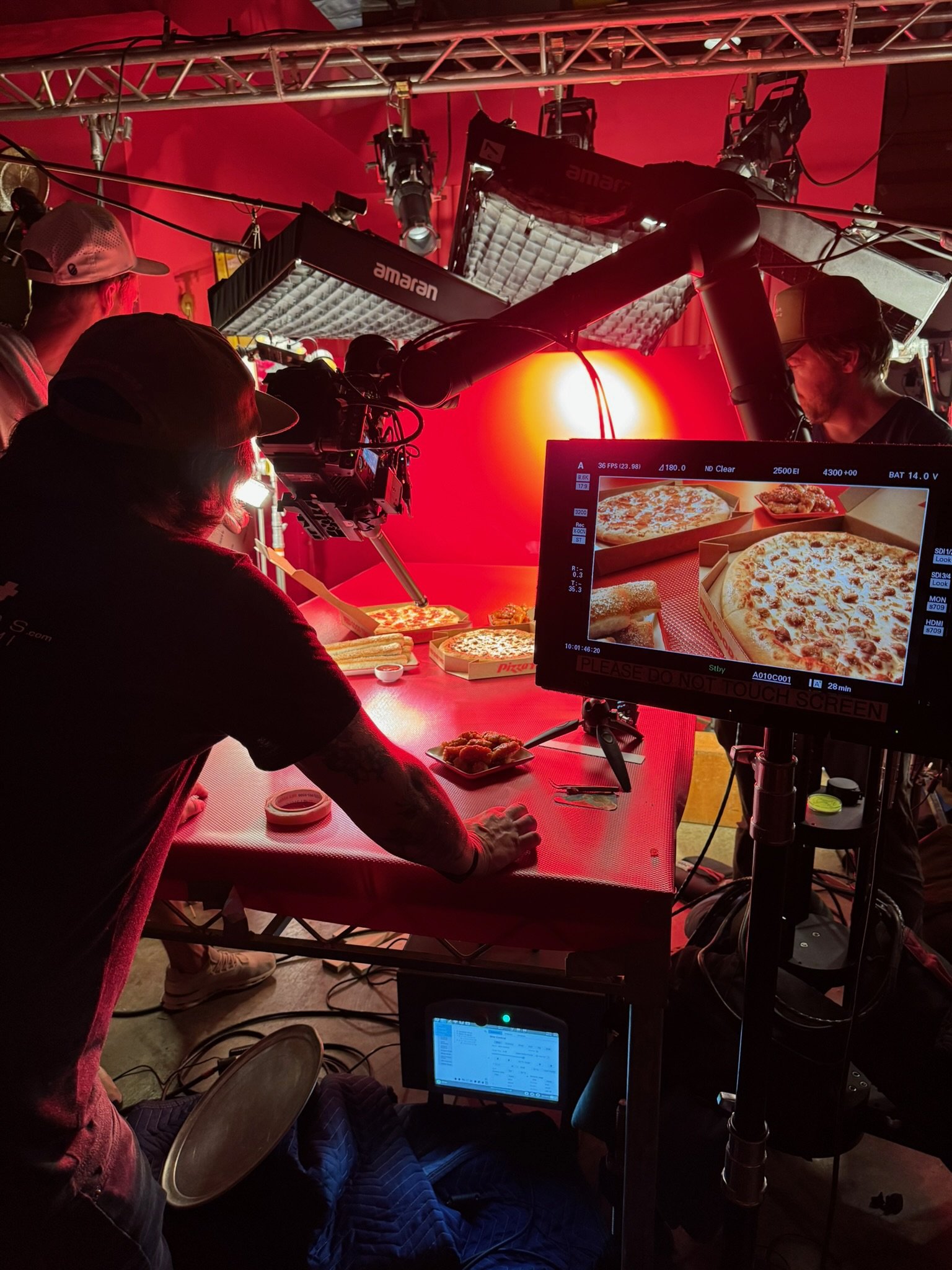 Behind the scenes of a pizza shoot, with multiple crew members operating camera and lighting equipment focused on pizza boxes and food on the table, with a monitor displaying the live view.