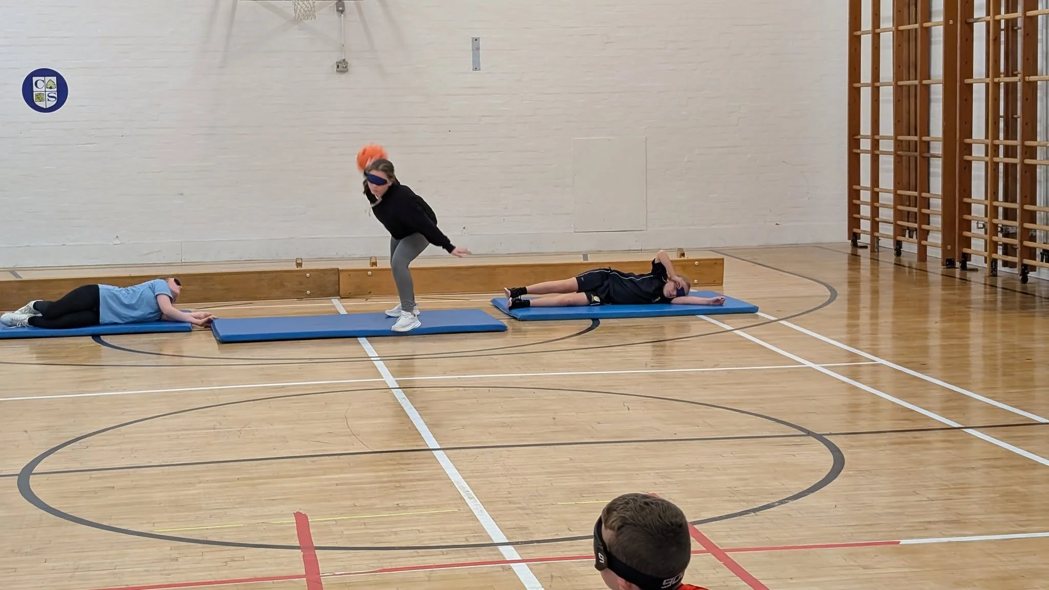 Two girls lying on gym mats, one girl in blue and one in black, with a woman in sportswear standing between them, in a gymnasium.