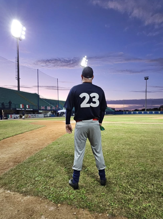 Baseball player standing on the field, wearing a navy jersey with the number 23, gray pants, and a black cap, with stadium lights and a evening sky in the background.