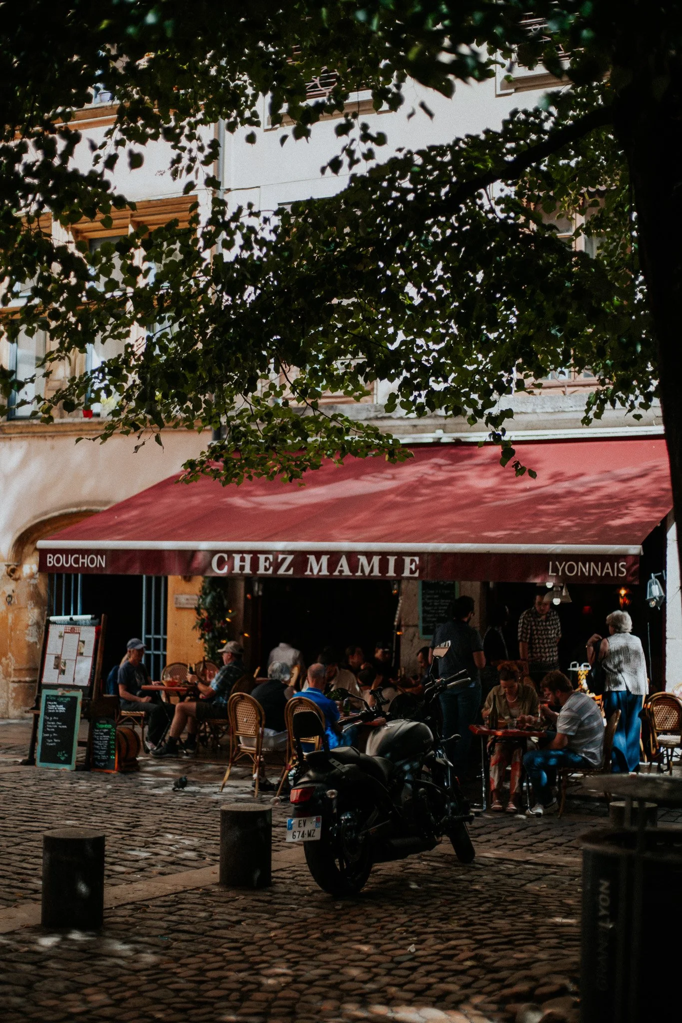 Outdoor seating at Chez Mamie restaurant under a red awning with several people dining and socializing, a parked motorcycle in the foreground, cobblestone street, and trees overhead.