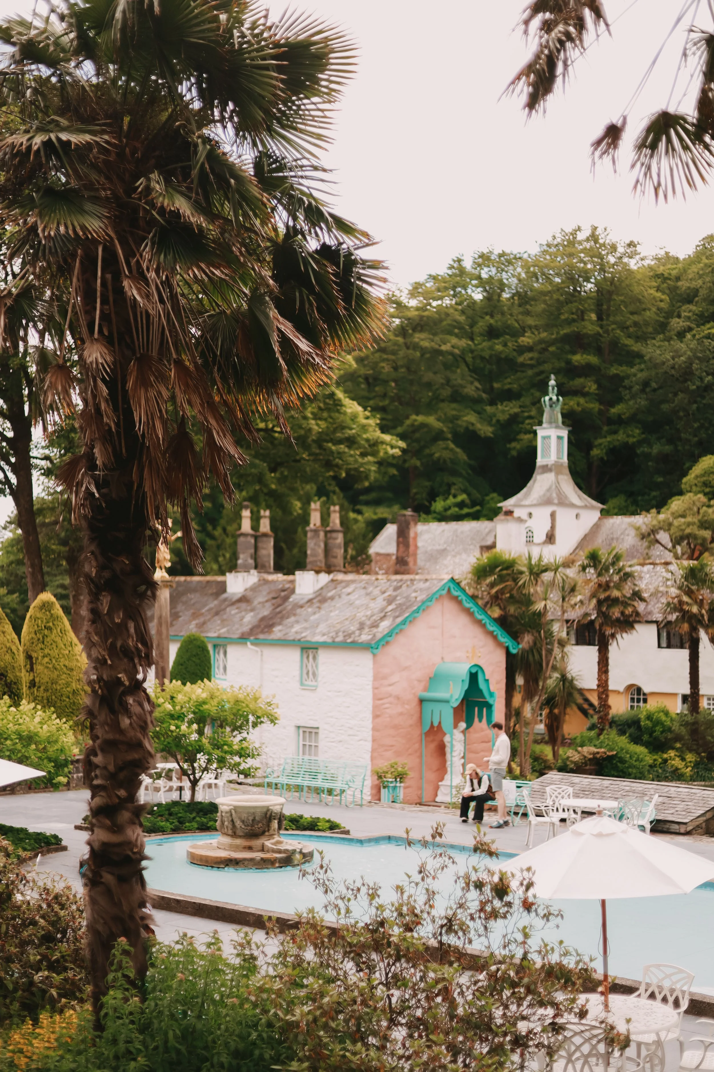 A scenic courtyard with a small fountain, surrounded by lush trees and colorful buildings, including a pink house with teal trim and a white building with a tower, under an overcast sky.