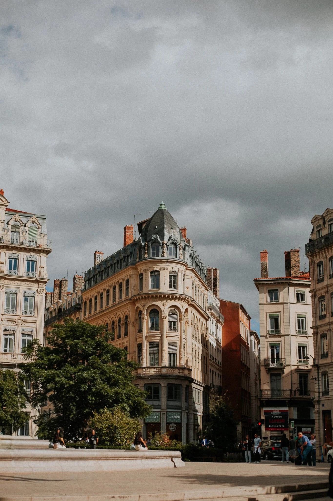 European city street with historic multi-story buildings, a tree, and people walking near a fountain, under a cloudy sky.