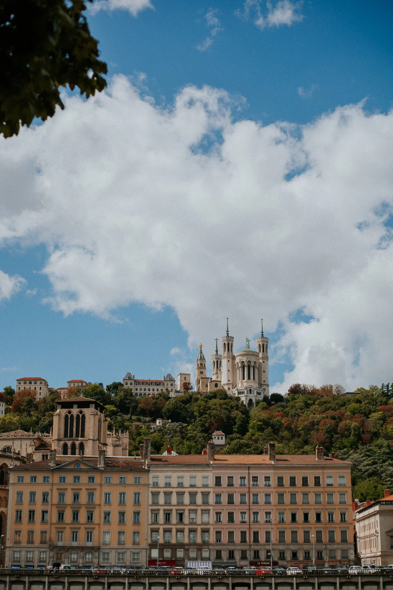 View of a hillside cityscape with historic buildings and the Basilica of Notre-Dame de Fourvière in Lyon, France, under a partly cloudy sky.