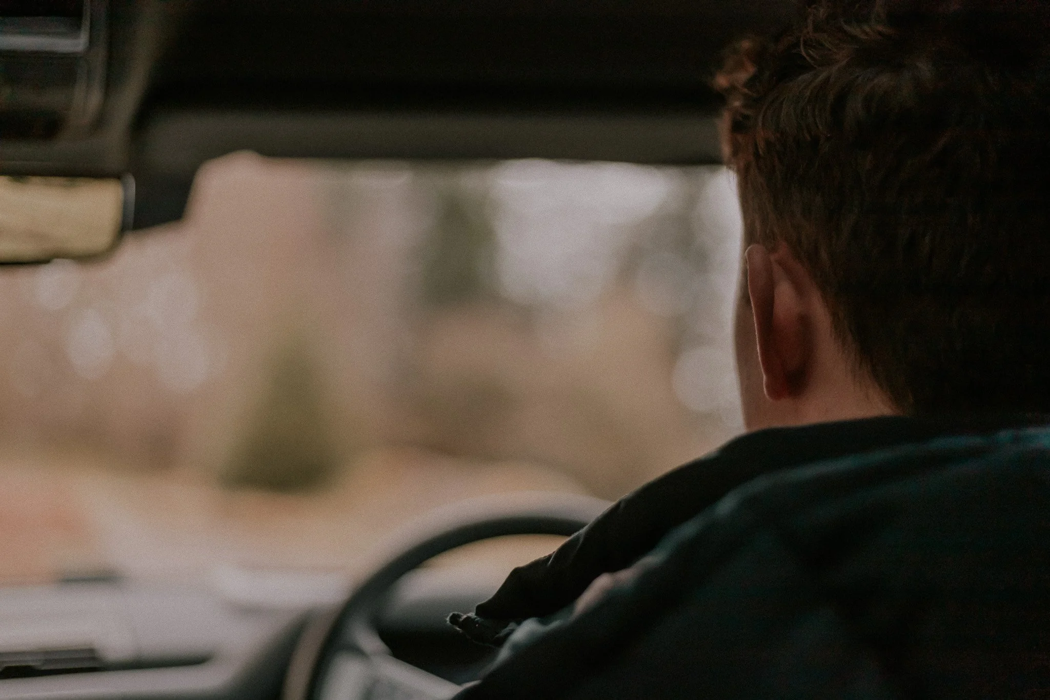 A person with short brown hair driving a vehicle, seen from behind, with a blurred outdoor background.
