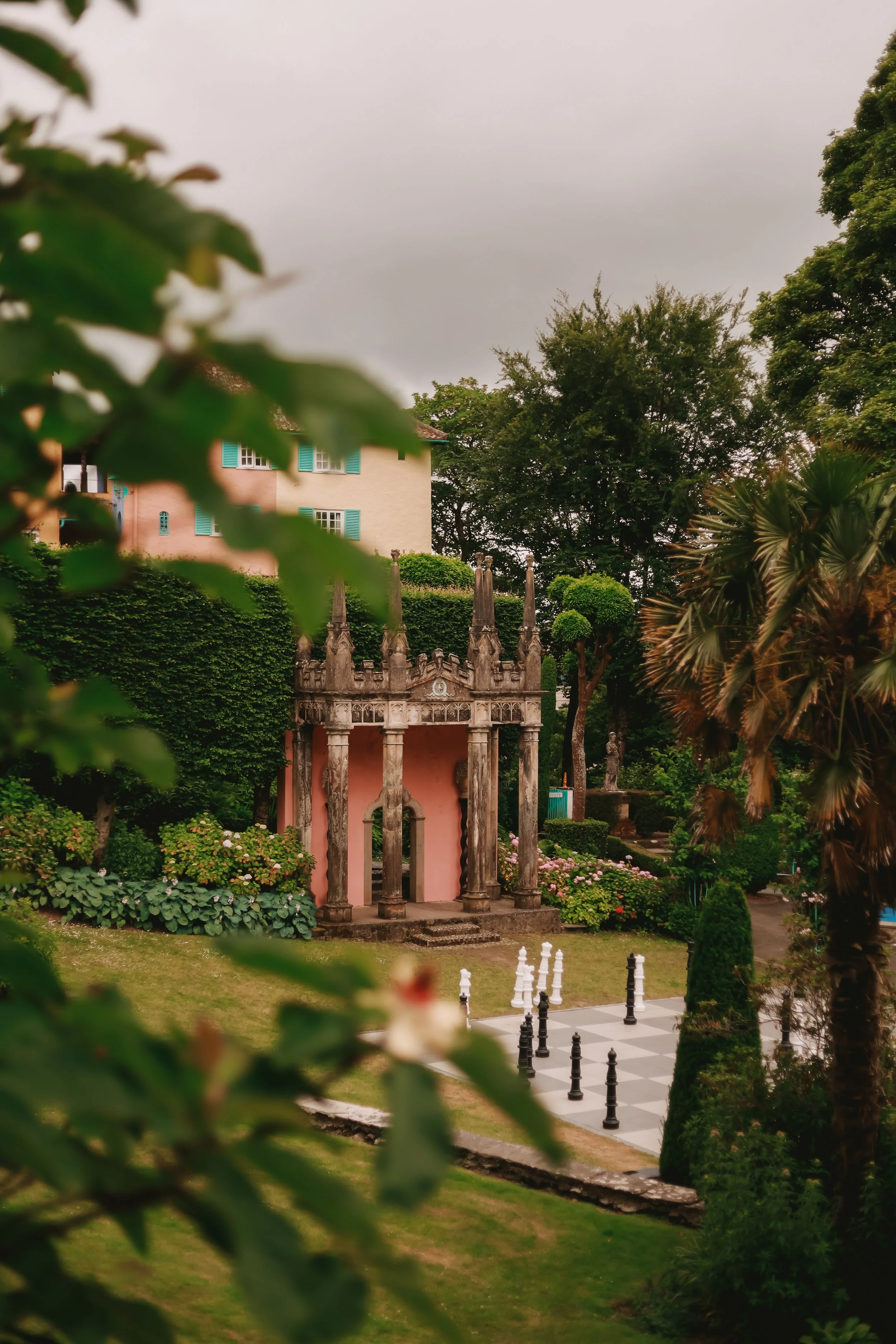 A garden scene with a classical stone archway, surrounded by lush green trees and plants, and an outdoor chessboard with black and white chess pieces.