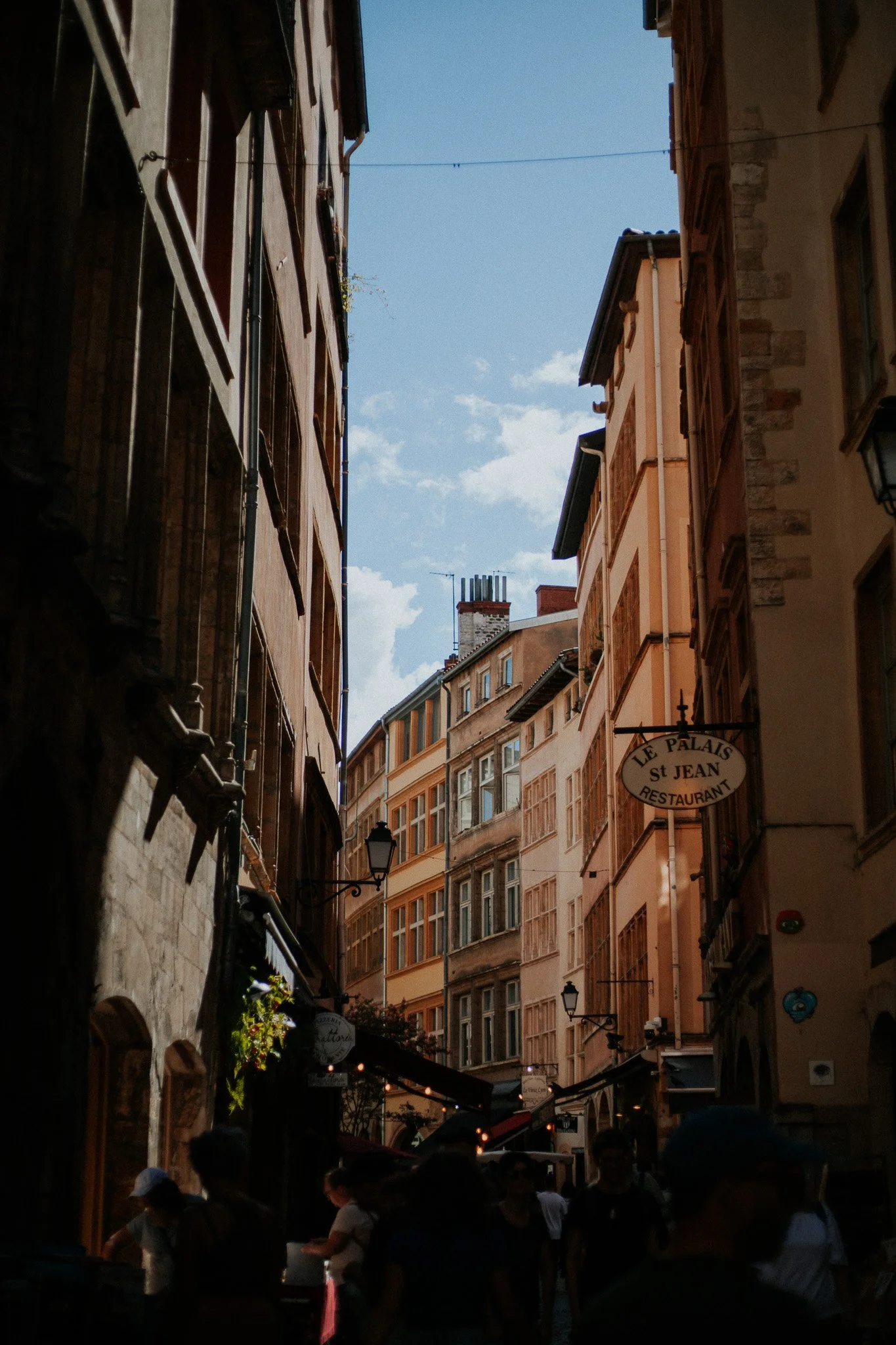 Narrow European street with tall buildings, people walking, and a sign for Le Palais St Jean restaurant.