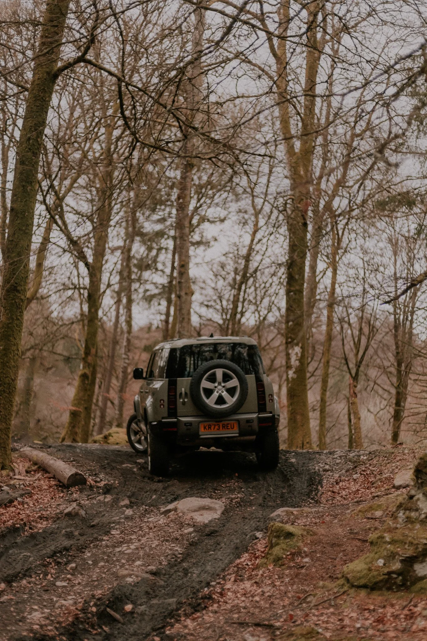 A rugged off-road vehicle driving through a dirt trail in a leafless forest during early spring or late fall.