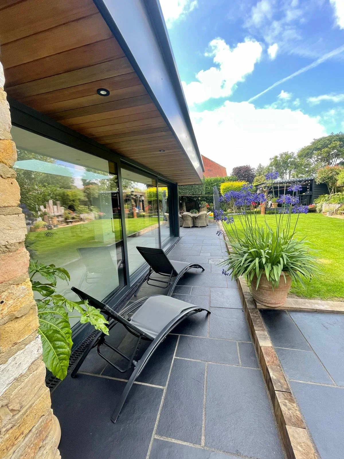 View of a patio with two lounge chairs next to a large glass door, overlooking a well-manicured garden with a large potted plant with purple flowers, a lawn, and outdoor seating under a sky with scattered clouds.