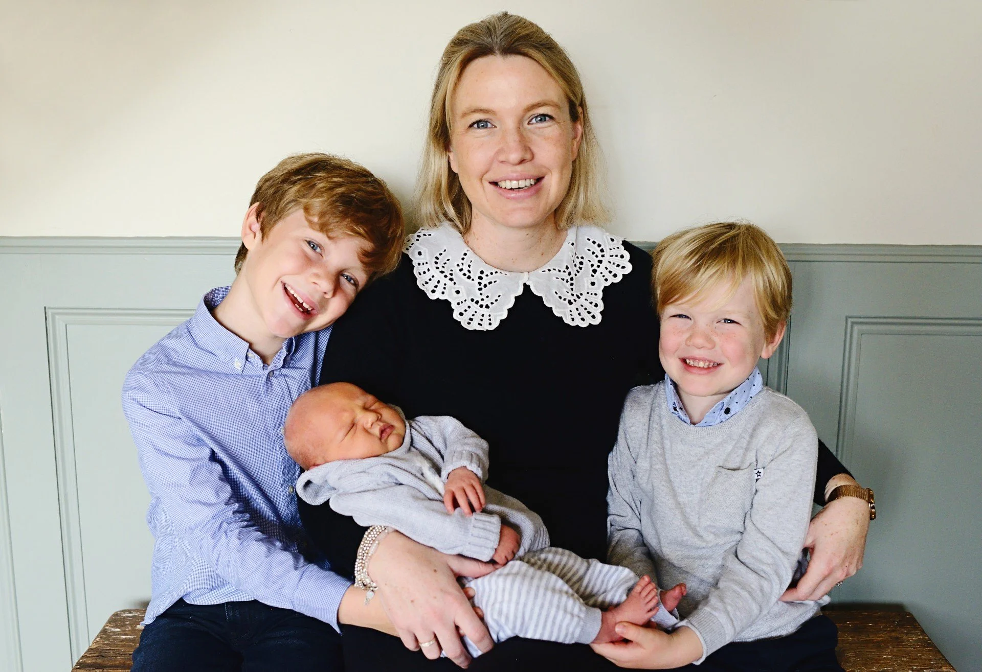 A woman sitting on a bench with four children, including a newborn, in a room with light green walls. The woman is smiling and wearing a black top with a white lace collar. The children are happily smiling; one boy leans on her shoulder, another boy is sitting beside her, and the baby is being held on her lap.