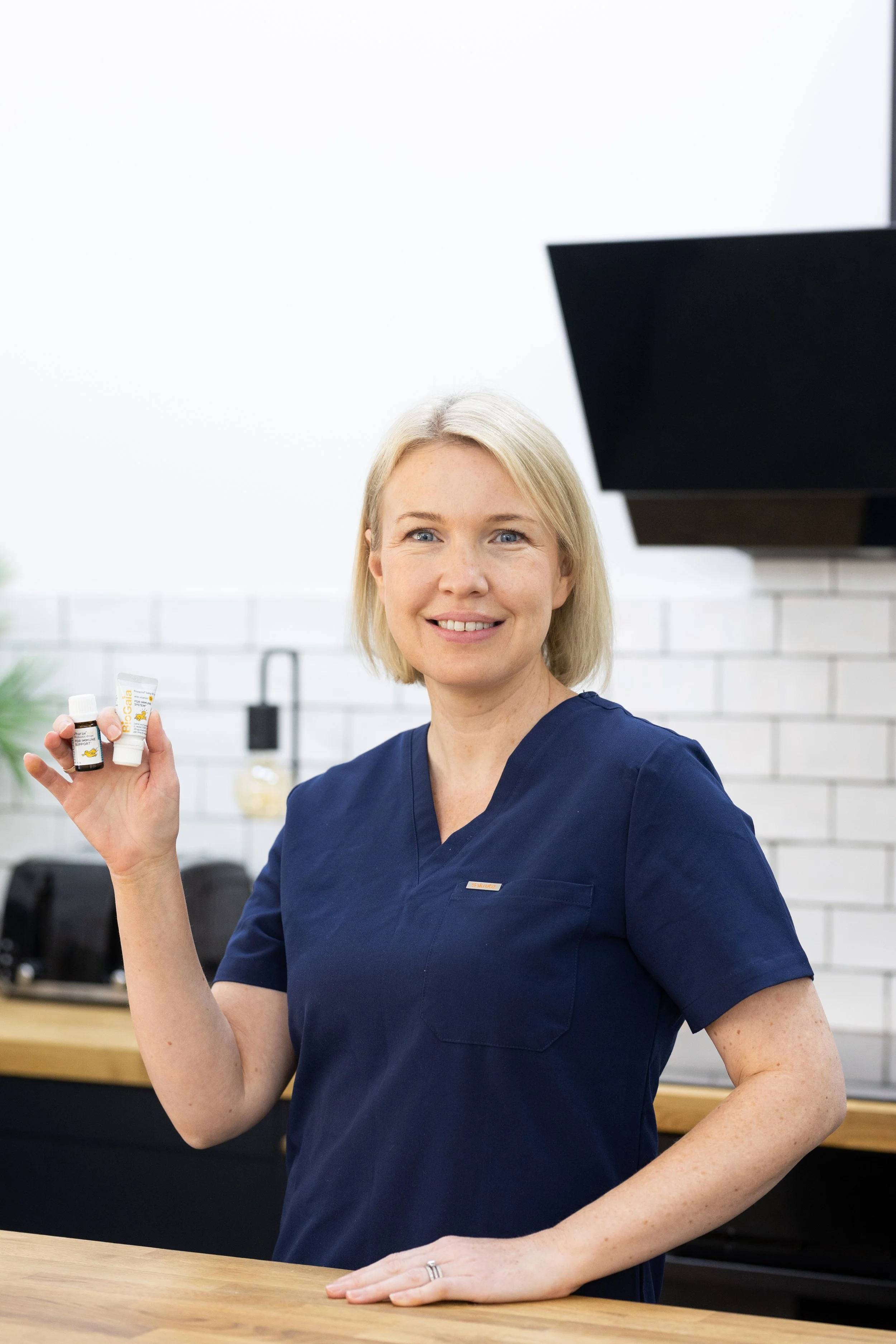 A woman with blonde hair wearing navy scrubs, smiling and holding small bottles of essential oils in a bright, modern kitchen.