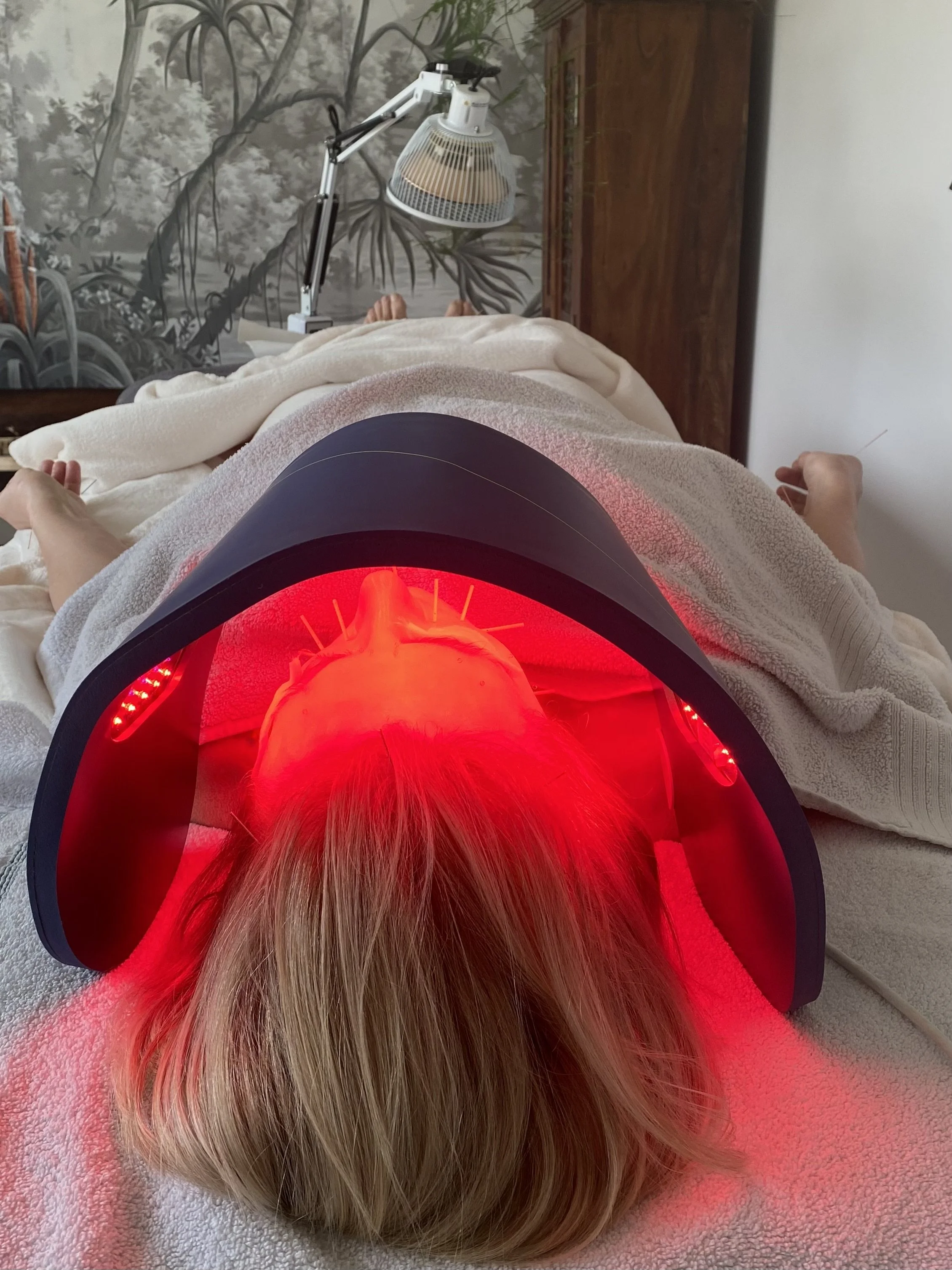 Person undergoing red light therapy on their face, lying on a bed with a blanket, with a lamp overhead emitting red light and a painting of trees and plants in the background.