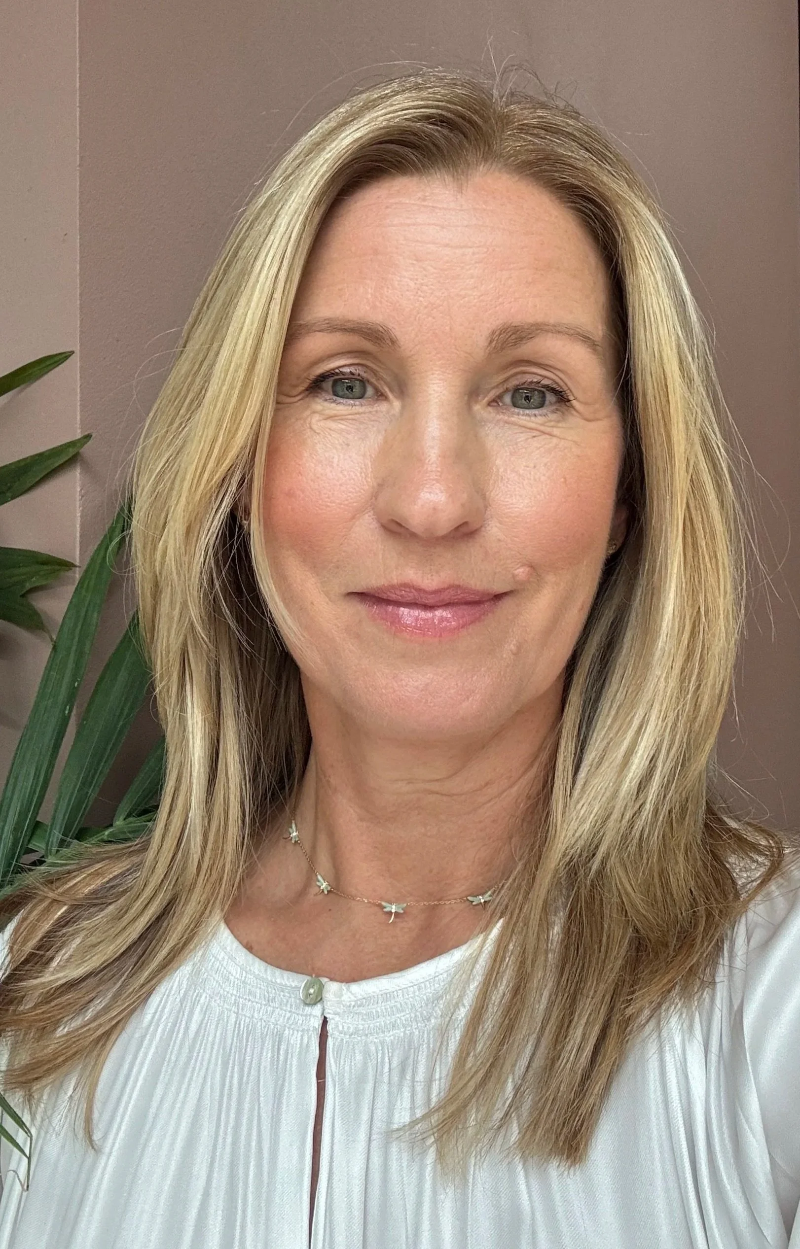 Close-up of a smiling woman with blonde hair wearing a white top and a delicate necklace with small charms, standing in front of a beige wall with a green plant visible on the left side.